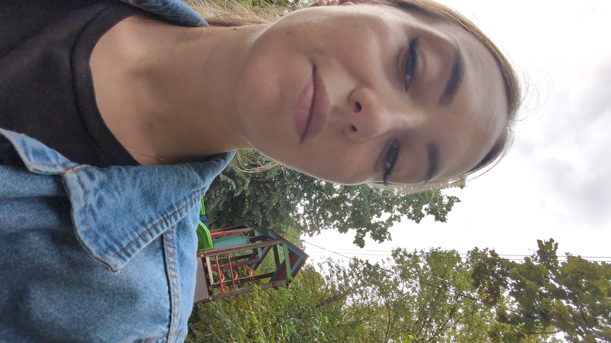A young woman, with light-colored hair and wearing a denim jacket over a dark t-shirt, is captured in a close-up, low-angle selfie. Her face is positioned towards the left side of the frame, with her eyes looking directly at the camera. In the background, a playground structure with red and blue accents is visible, partially obscured by the lush green foliage of trees. The sky above is overcast with clouds. The scene appears to be outdoors, possibly in a park or public space in Chișinău, Moldova.