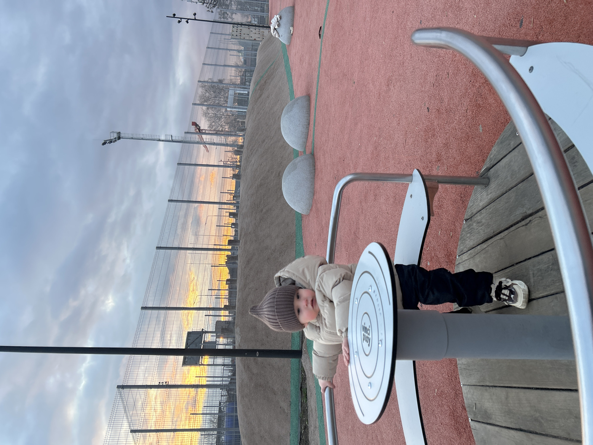 A baby wearing a tan winter hat and coat and black pants with white sneakers is playing on playground equipment. The equipment is a circular platform with metal bars. The playground has a red rubber surface. In the background, there is a fence and a construction crane in the city of Klin, Russia. The sky is overcast with hints of orange light.