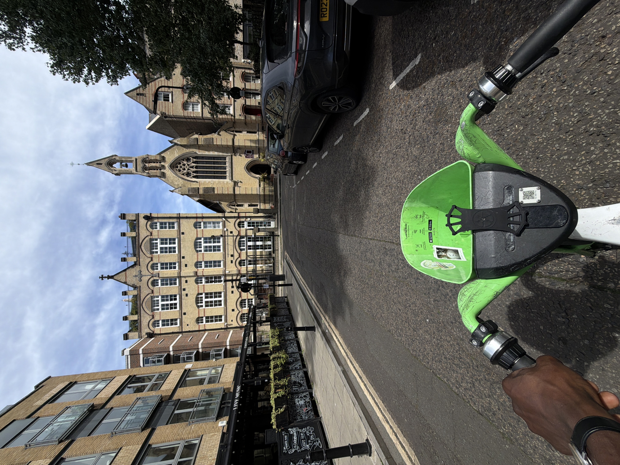 The image depicts a street scene in the city of London, United Kingdom. A first-person perspective shows a person's hands gripping the handlebars of a bright green electric scooter. The scooter's front basket is visible, with some stickers and instructions. The street is paved and lined with yellow double lines indicating no overtaking. On the left side of the street, a modern brick building with multiple floors and balconies is prominent, with a black awning and signage that reads "LIVE MUSIC". Adjacent to this building is a row of planters with greenery. Further down the street, a historic yellow brick church with a distinctive bell tower and arched windows stands, indicating a mix of old and new architecture. Several cars are parked on the right side of the street, including a dark grey SUV with a license plate starting with "RO23". The sky above is partly cloudy. The overall impression is of a sunny day in an urban environment.