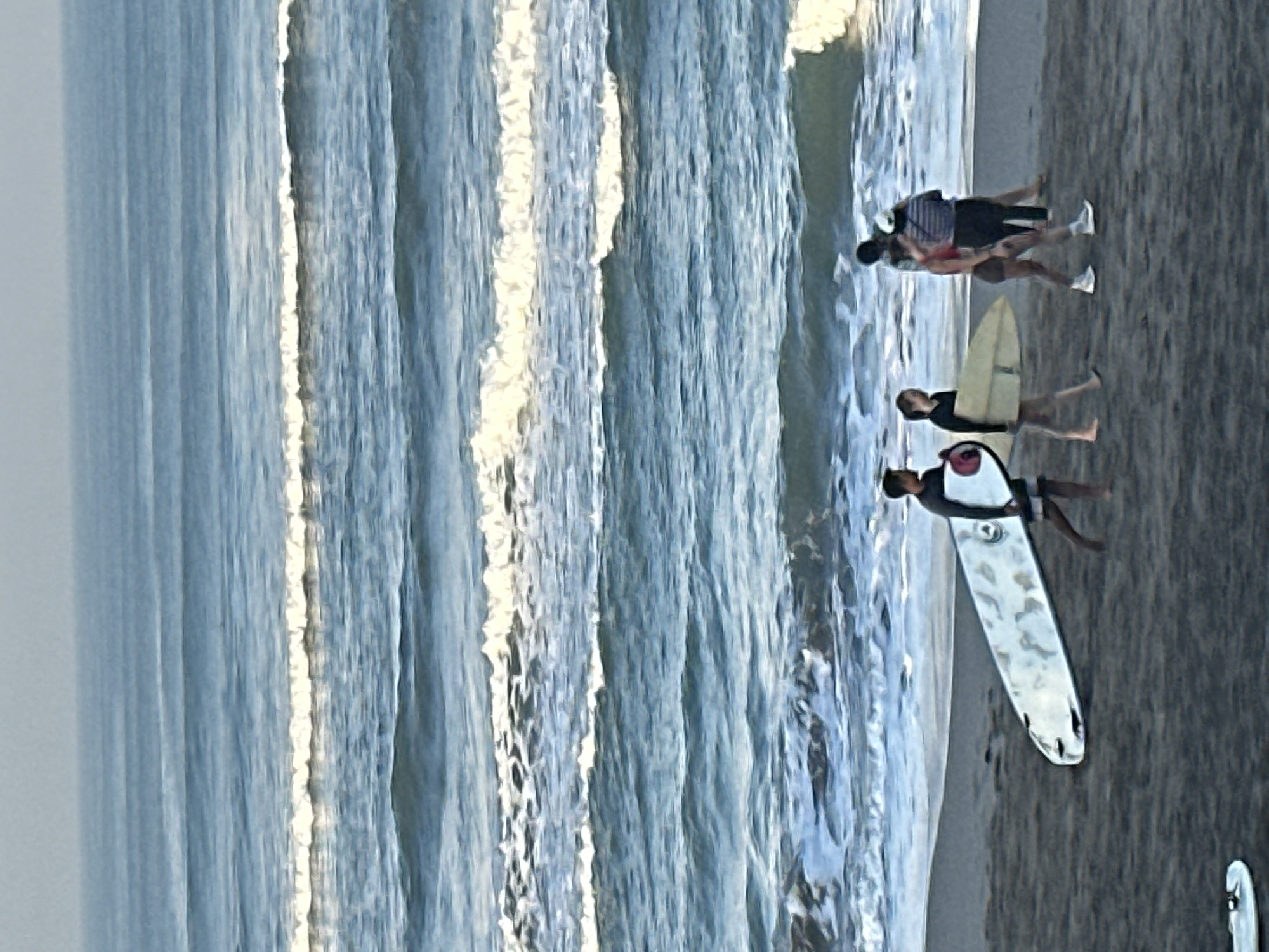 A group of four people walk along the shore of a beach in North Kuta, Indonesia. The ocean is to the left of the frame, with several lines of blue waves and white foam. The beach is dark gray sand and occupies the bottom half of the frame. The people are walking from left to right, with two people in front and two people behind them. The person on the far left is a man carrying a white surfboard. Next to him is a woman holding a yellow surfboard. Behind them, a man and a woman walk together, with the woman in front. The water is calm and clear. The sky is a pale blue and hazy.