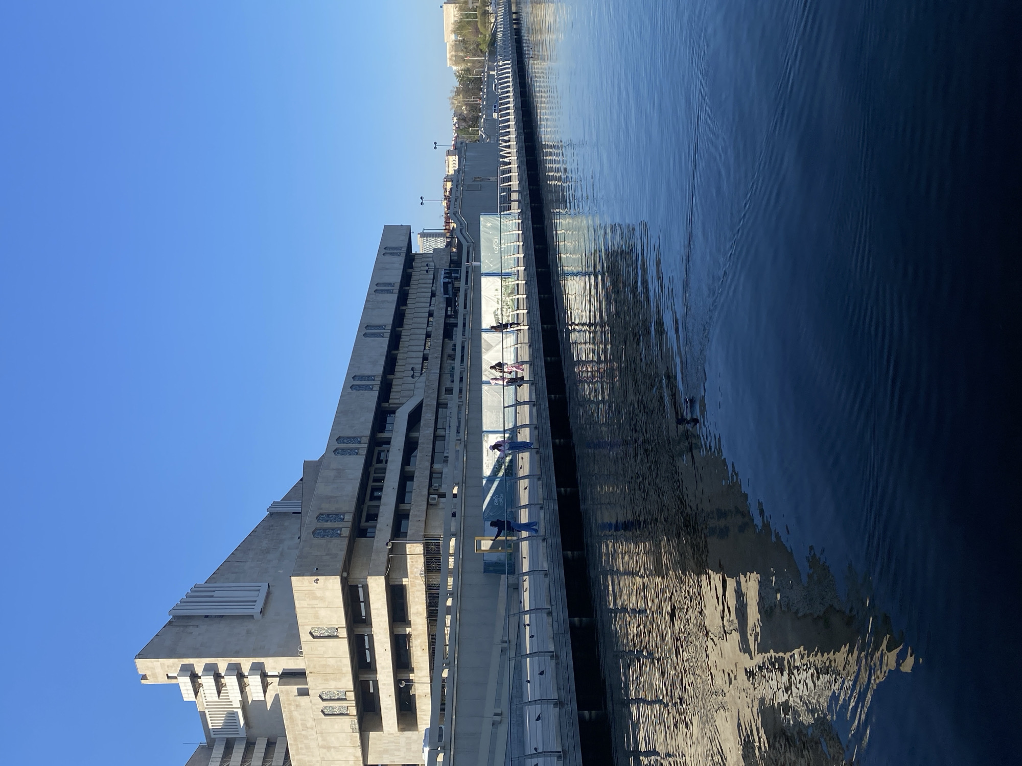 The image shows the Ministry of Agriculture and Food of the Republic of Tatarstan in Kazan, Russia. The building is a large, modern concrete structure with a unique and somewhat brutalist design. It is situated next to a body of water, possibly the Kazanka River. People are walking along a path or boardwalk that runs alongside the building and the water. The sky is clear and blue. The water reflects the building and the sky.