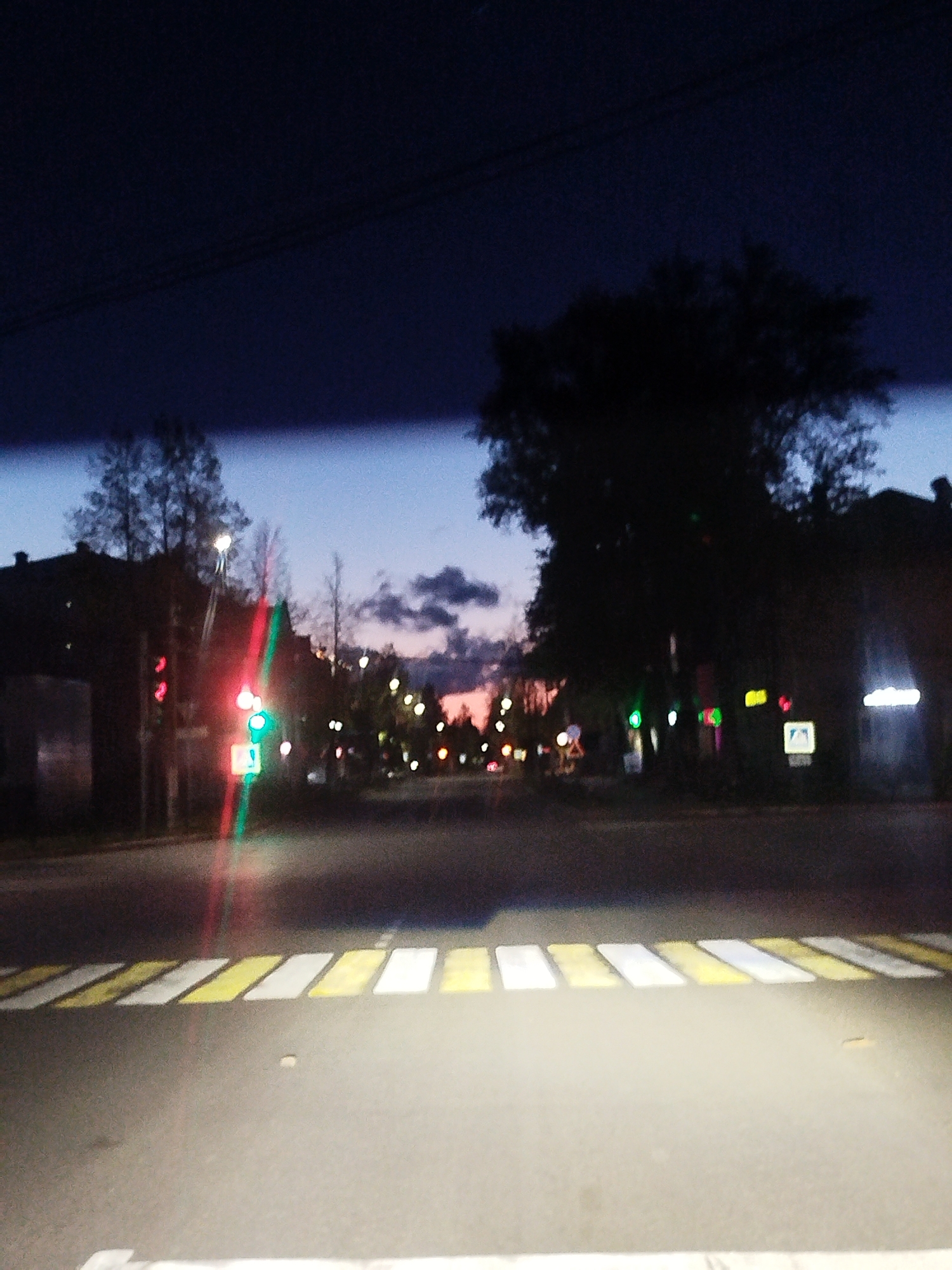 The image shows a street scene at dusk in Chaikovsky, Russia. The photograph appears to be taken from a vehicle, as a portion of the vehicle's interior is visible at the top. A pedestrian crosswalk is in the foreground. Across the street, traffic lights are visible, with one showing red and another showing green. Buildings and trees line the street. In the background, the sky shows the colors of sunset. Streetlights and signs are visible along the street.