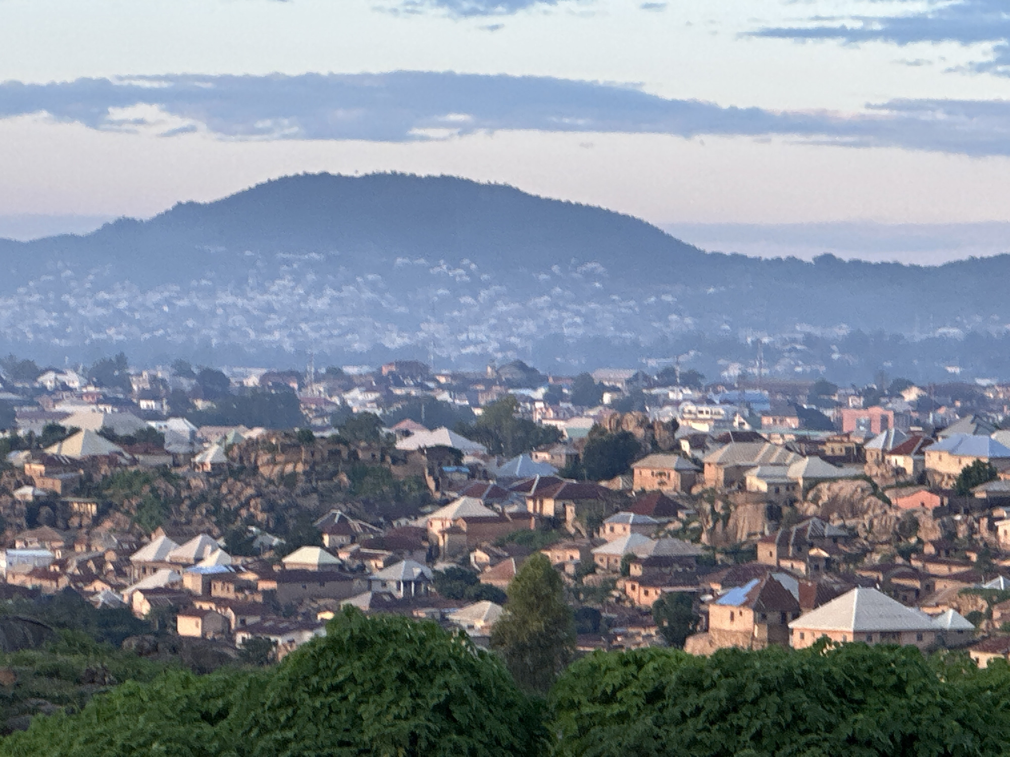 The image shows a panoramic view of a dense urban landscape on a hillside in Jos, Nigeria. The scene is characterized by a multitude of buildings with varied roofing materials and architectural styles, tightly packed together on the uneven terrain. Rocky outcrops are interspersed with the structures, suggesting a challenging topography. In the foreground, lush green foliage forms a natural border, obscuring the lower portion of the settlement. In the background, a hazy, mountainous silhouette rises, with a lighter, more diffuse settlement visible on its slopes. The sky above is a soft gradient of pale blue and white, with subtle cloud formations suggesting either early morning or late afternoon light. There are no visible people, actions, or interactions in the image. The overall impression is one of a sprawling, established community integrated into its natural environment.