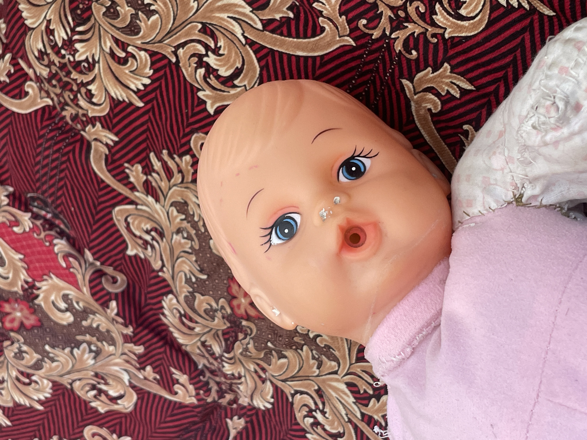The image shows a close-up, overhead view of a doll lying on a patterned fabric. The doll has an open mouth, blue eyes, and is wearing a pink garment. The fabric it rests on features a dark red background with black stripes and a repeating ornate brown and gold pattern. The image appears to be taken indoors, possibly in a residential setting. There is no visible indication of location in Tashkent, Uzbekistan.