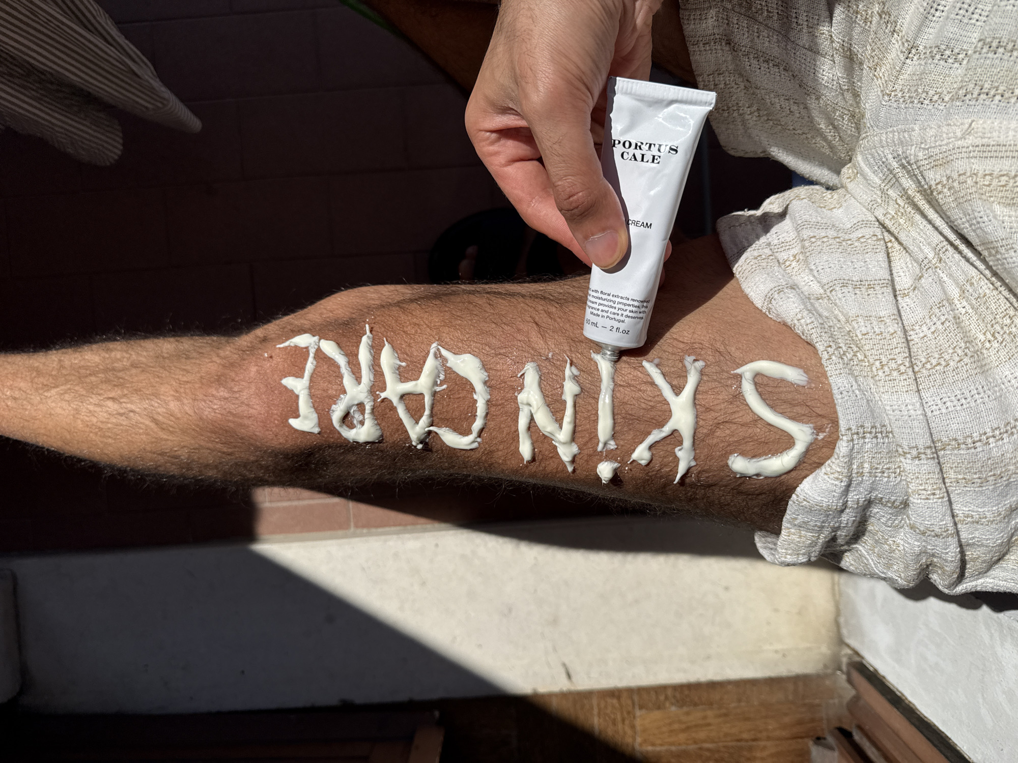 A man is applying white cream from a tube labeled "PORTUS CALE CREAM" onto his hairy leg, writing "SKIN CARE." The background includes a tile floor and wall, partially visible. The scene is well-lit, presumably indoors. The location in Milan, Italy is not visually discernible from the image.
