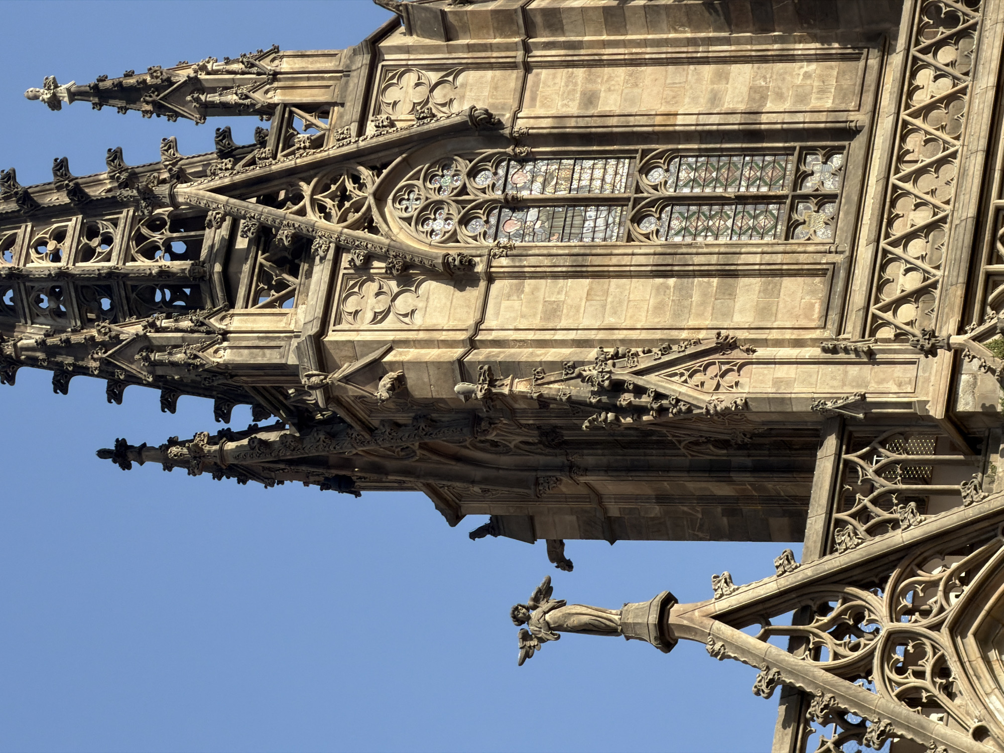 The image shows a close-up of a section of the exterior of the Barcelona Cathedral, a Gothic Roman Catholic cathedral located in the heart of the Barri Gòtic (Gothic Quarter) of Barcelona, Spain. The focus is on the intricate stonework of the cathedral's spires and facade, under a clear blue sky.
Prominently featured is a statue of an angel with outstretched wings, standing atop a pointed spire. The angel is sculpted from stone and appears to be holding something in its hands, possibly a scroll or a trumpet, though the detail is softened by distance and the material. To the right of the angel, and slightly behind it, is a gargoyle, a common architectural feature on Gothic cathedrals, which serves as a water spout.
The main body of the cathedral visible is a tall, ornate spire characterized by complex Gothic tracery. This includes multiple pointed arches, decorative fenestration with quatrefoil and trefoil motifs, and smaller pinnacles. One large stained-glass window with multiple lancets is visible within the main spire. The glass is multi-colored and depicts figures, suggesting it is a religious scene. The stonework itself is weathered and a warm, sandy color, indicating age.
Below the main spire, further decorative elements are visible, including a latticework frieze and other pointed arch structures, all rendered in the same Gothic architectural style. The overall impression is one of elaborate and detailed craftsmanship, typical of medieval European cathedrals. The lighting suggests it is daytime, with sunlight illuminating the stone and casting some shadows, highlighting the three-dimensionality of the architectural details. The clear sky indicates fair weather.