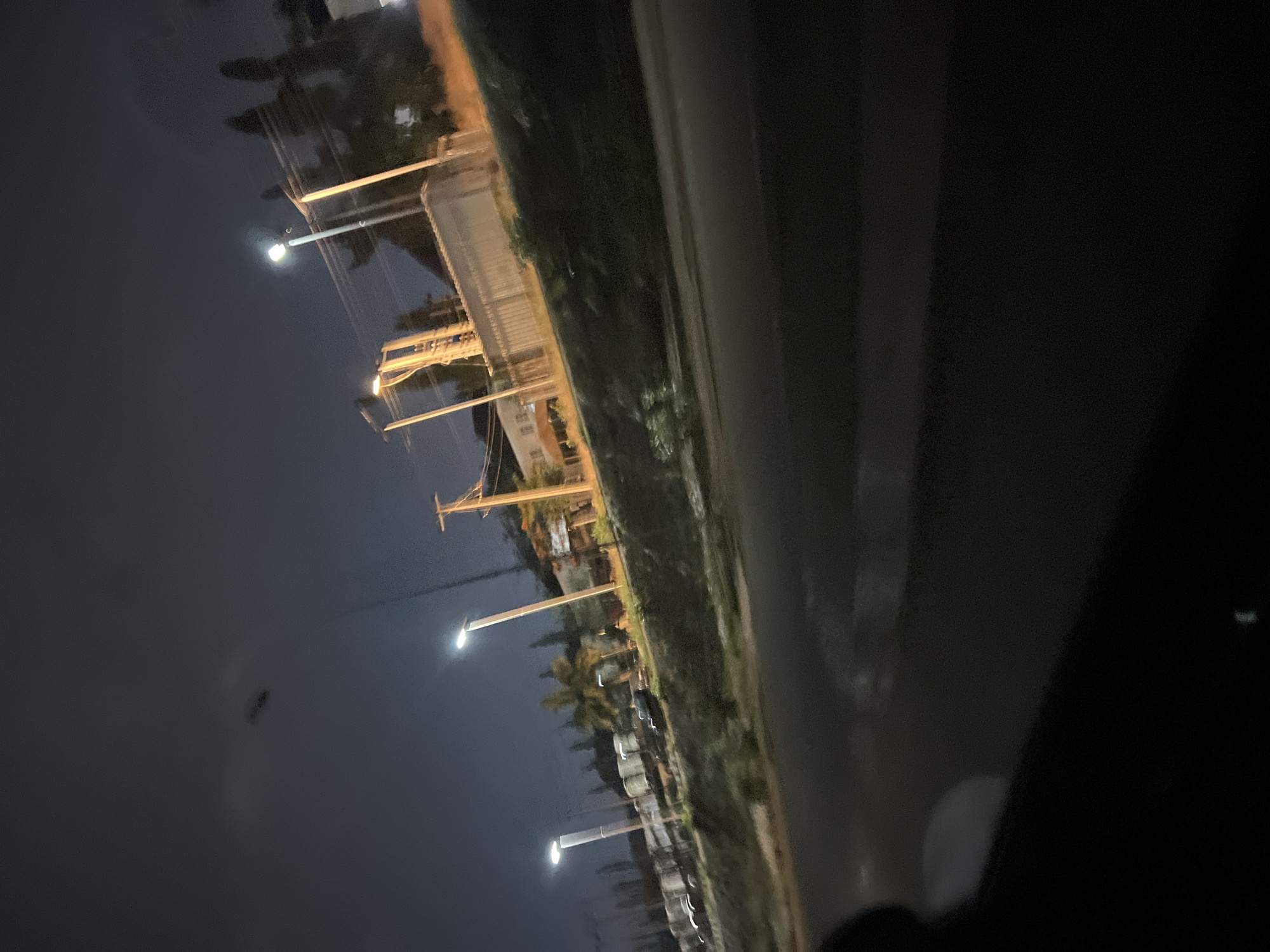 The image depicts an urban street scene at night, potentially in or around Jiru, Nigeria. A row of streetlights illuminates a road bordered by vegetation on one side and what appears to be buildings further back. Utility poles with wires are visible, suggesting an infrastructure for electricity and communication. The image is taken from the perspective of being inside a vehicle. The light is dim, typical of nighttime conditions in a city.