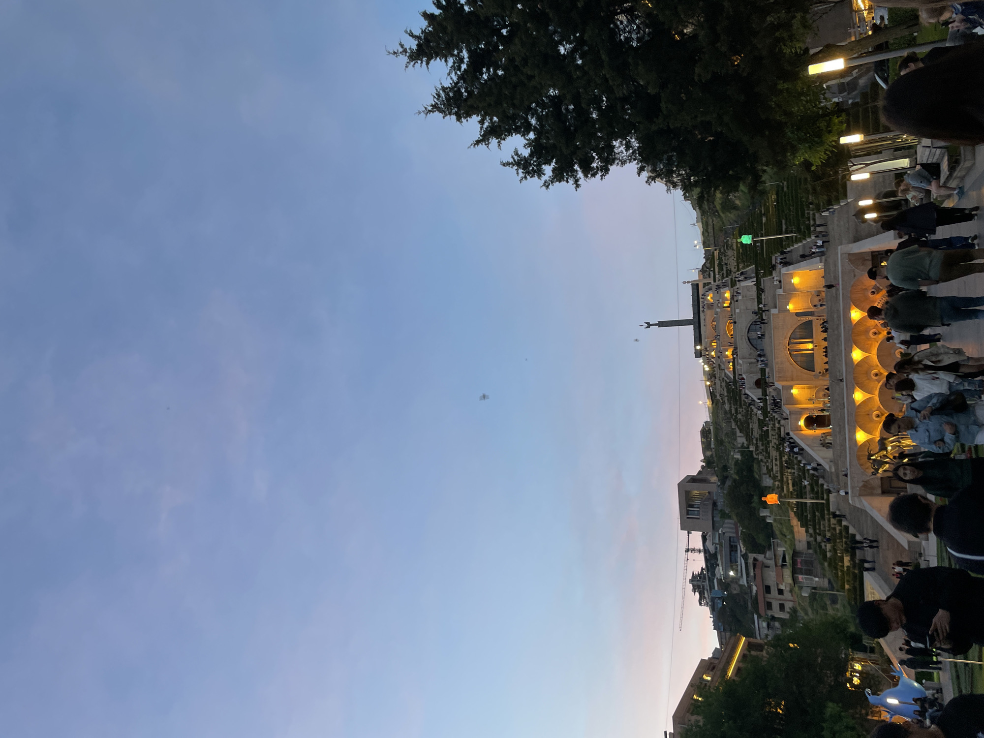 The image shows the Yerevan Cascade in Armenia at dusk. The Cascade is a giant stairway adorned with sculptures, fountains, and gardens. People are walking up and down the stairs. The architecture is illuminated with warm lighting. The sky is a gradient of blue and pink. A tree is visible in the upper left corner. Buildings and a construction crane are visible in the distance behind the Cascade.