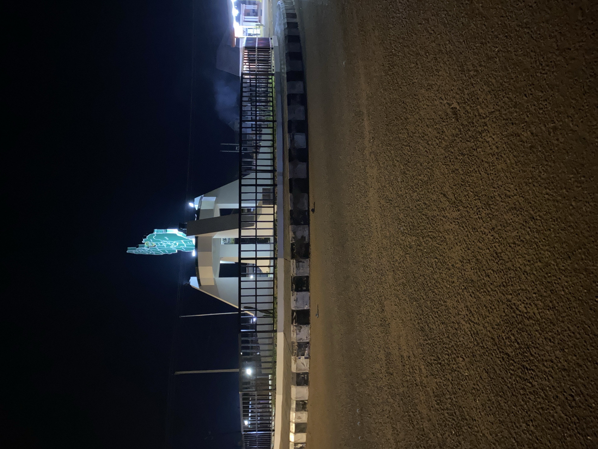 The image depicts a night scene in Bukuru, Nigeria. A lit, green and white map of Plateau State sits atop a structure with white columns and a dark interior. A black metal fence runs along the edge of the structure, adjacent to a road. The road surface appears to be asphalt. Streetlights are visible in the background. The sky is dark. There are no people visible.