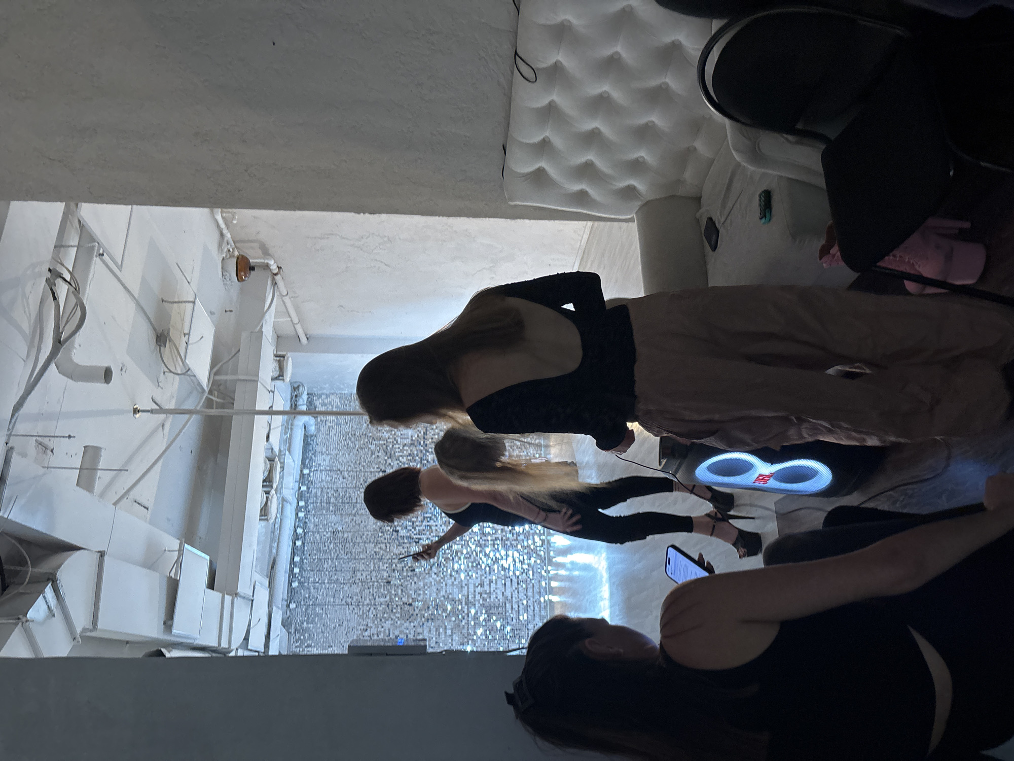 The image shows a dimly lit, modern interior. In the foreground, a person in black is holding a phone. Behind them, a group of four women are visible, some of whom are looking at their phones. One woman, in the center, is wearing a black top and black pants, and her hair is long and blonde. Another woman, to her left, has dark hair and is also wearing black. A third woman, on the far right, has long brown hair and is wearing a black top and beige pants. She appears to be posing. In the background, there is a white tufted sofa and some architectural elements that suggest an industrial or minimalist design. A large speaker with illuminated circular elements is visible on the floor. The overall impression is one of a gathering or a photoshoot. The time of day is not discernible, and there are no obvious weather cues. The location appears to be indoors, possibly a studio or a chic lounge. The mood is casual yet posed. There is no visible text in the image.