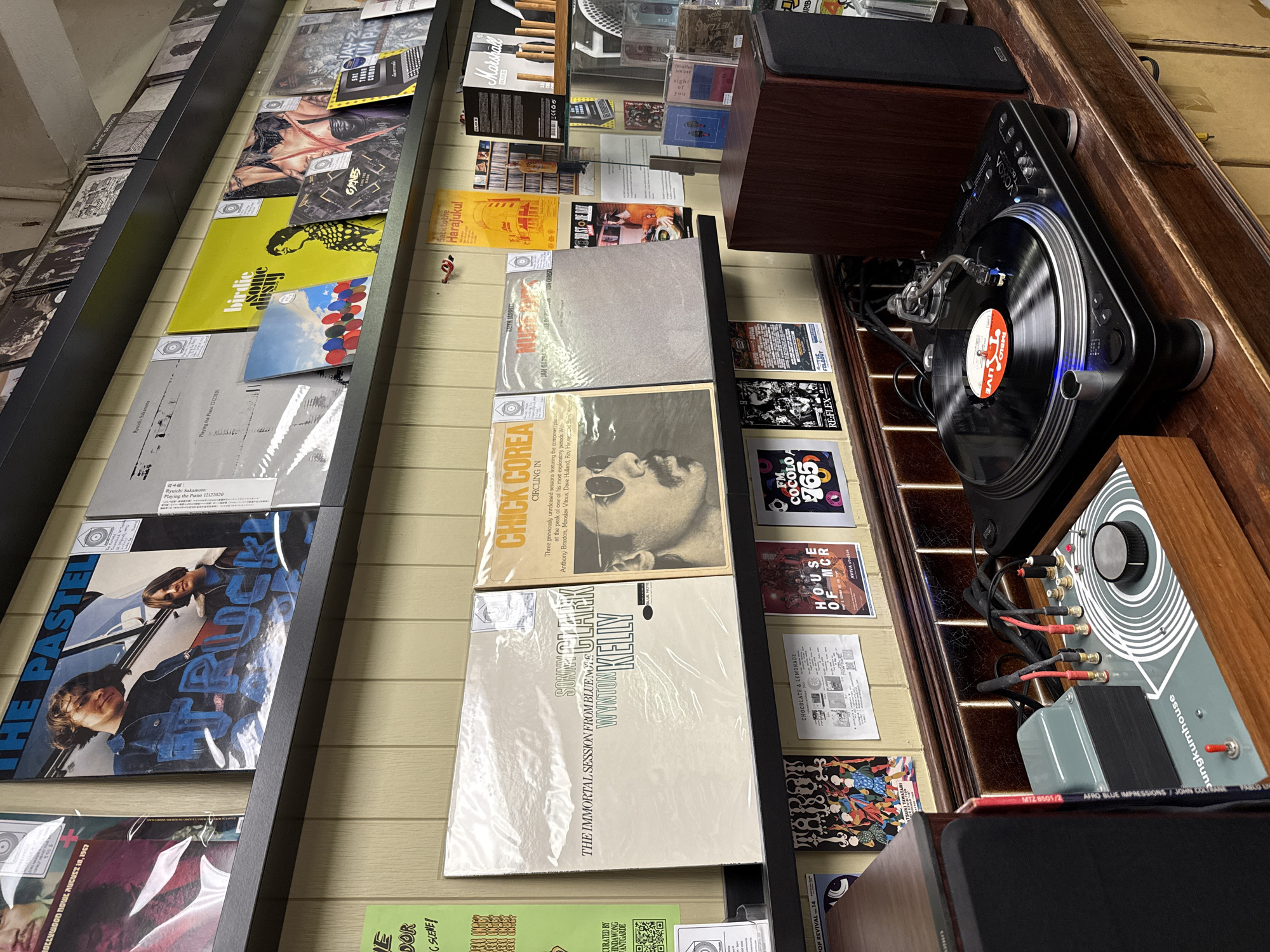 A cluttered record store shelf displays vinyl records and audio equipment.
Vinyl records are stacked on a black shelf, with covers facing forward. Record titles include "THE PASTELS", "Ryuichi Sakamoto: Playing the Piano", "THE IMMORTAL SESSION FROM BLUE NOTE", "SONNY CLARK", "CHICK COREA", and "CIRCLING IN".
On the right, a turntable with a black base and silver accents is playing a record with a red label. Next to the turntable is a wooden audio amplifier with a control panel. Cables connect the turntable to the amplifier.
The wall behind the shelf is decorated with small posters and advertisements, including mentions of "HOUSE OF MCR AVIVA studios", "FM COCOLO 765", "REFLEXLDOUT", "TAROT", and "birdie hisap some".
The location is hinted to be in Bangkok, Thailand, through a poster with Thai script and the phrase "CURATED BY HINDAWONG ANTGARDE".