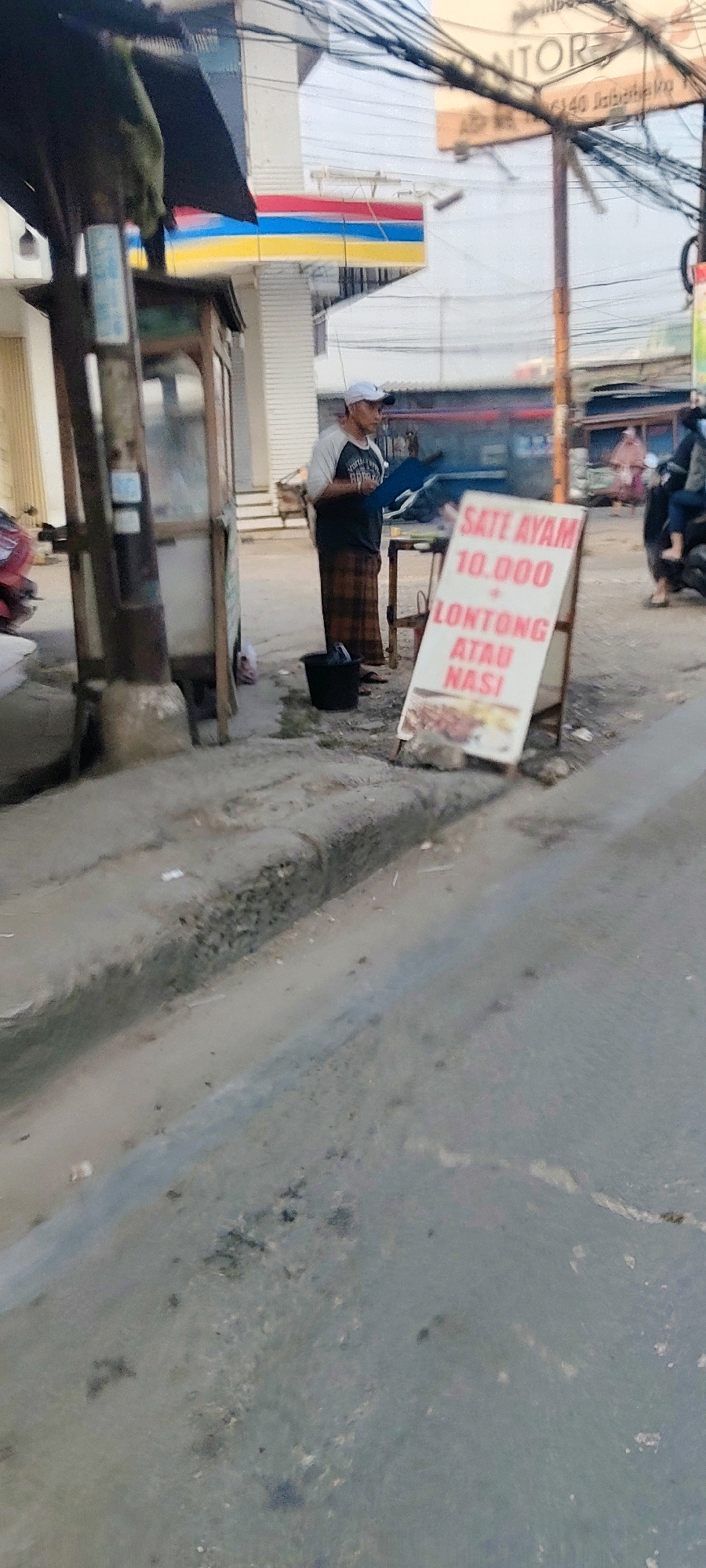 The image depicts a street scene in Mekarmukti, Indonesia. In the foreground, a portion of a street and sidewalk is visible, with some debris and uneven surfaces. A man, wearing a white cap, a dark t-shirt with a graphic, and a checkered sarong, stands near a food stall. The food stall has a dark wooden structure with a glass display case. A black bucket is on the ground near the man.
To the right of the man, an A-frame sign displays text in red. The sign reads "SATE AYAM 10.000 LONTONG ATAU NASI". Below this text, there appears to be an image of sate ayam.
In the background, a building with a prominent horizontal stripe of blue, red, and yellow is visible, likely a commercial establishment. Overhead, a tangle of electrical wires crisscrosses the sky. Further in the background, other buildings and a motorcycle with a passenger are dimly discernible. The overall lighting suggests it is daytime, possibly late afternoon.