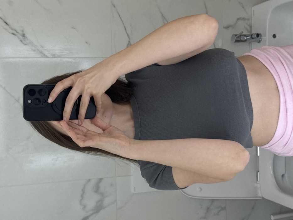 The image depicts a mirror selfie taken by an individual in a bathroom located in the city of Scafati, Italy. The subject, an individual with visible dark brown hair, is wearing a dark grey, short-sleeved, ribbed crop top and light pink bottoms. Both hands are holding a black smartphone with a multi-lens camera array and a circular MagSafe ring on its back, obscuring the upper portion of the individual's face. The bathroom features white marble-effect wall tiles with grey veining. In the background, a white toilet with an open lid is visible to the left, and a white bidet with a chrome faucet is visible to the right.