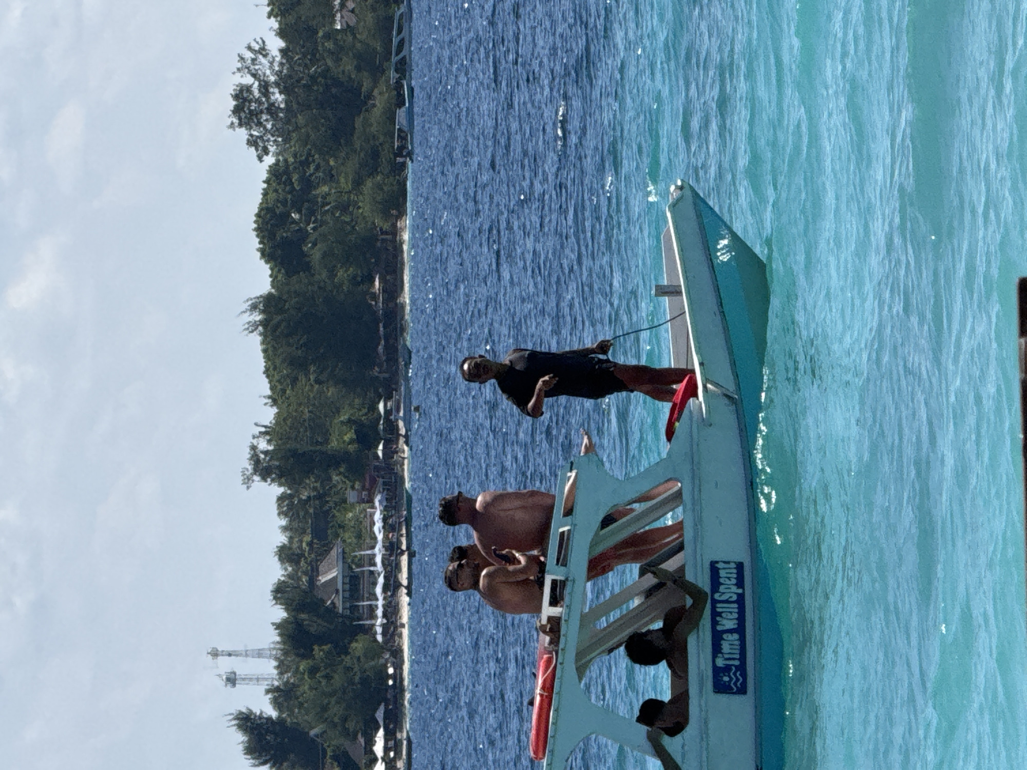 The image shows a group of people on a boat in the water off the coast of what appears to be Unknown, Indonesia. The boat has the words "Time Well Spent" written on its side. The water is a clear turquoise color, and the coastline in the background includes vegetation and buildings. One person is standing at the front of the boat. Several other people are sitting or standing on the boat.
