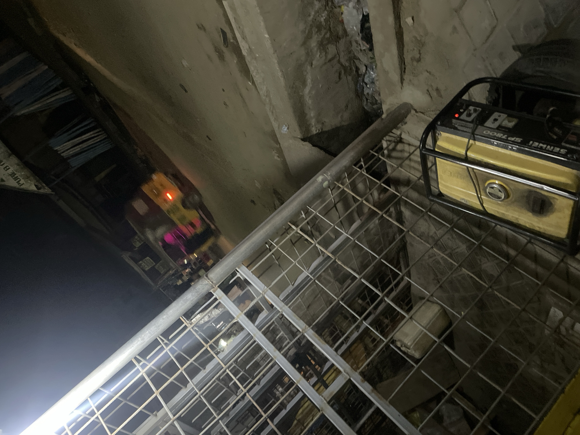 A low-angle, underexposed image shows a portable generator placed on a metal grid structure, possibly a staircase or elevated platform. The generator is yellow and black, with visible controls and markings. The grid is made of interconnected metal bars forming square or rectangular patterns. In the background, a poorly lit street scene is visible, with blurry lights suggesting vehicles or buildings. The surrounding environment appears to be an urban, possibly industrial, setting, with concrete structures. The location is Unknown, Unknown. The general atmosphere is dim and somewhat gritty.