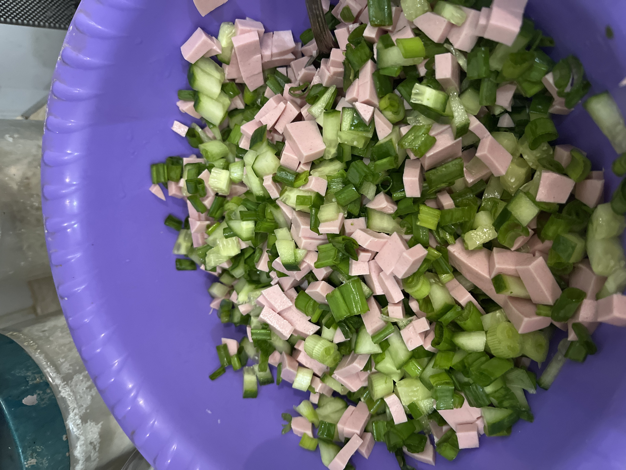 A vibrant purple plastic bowl, seen from a top-down perspective, is filled with a mixture of diced ingredients, indicative of food preparation in a kitchen. The main components are uniformly cut, light pink cuboid pieces, consistent with cooked ham or sausage, intermingled with two types of green vegetables. These include lighter green, somewhat translucent diced pieces, likely cucumber, and darker green, ring-shaped or cylindrical pieces, identifiable as chopped green onions or scallions. The ingredients are thoroughly mixed within the bowl, suggesting they are being combined for a salad or similar dish. A metallic utensil, possibly a spoon or part of a knife, is subtly visible among the ingredients, implying active preparation or serving. The background offers glimpses of a light-colored surface, presumably a countertop, and a partial view of another container, possibly blue or teal, with some white residue on it. There are no people visible, nor is there any discernible text. The image captures a factual and straightforward moment of culinary activity.