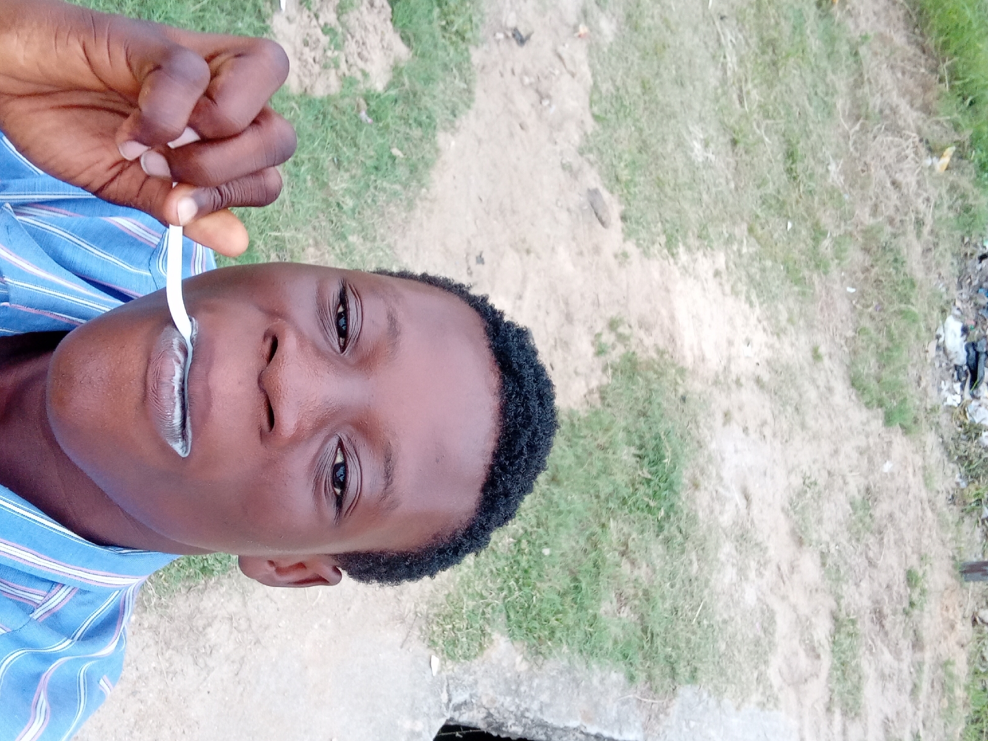 A young Black person is taking a close-up selfie outdoors. They have short, dark, curly hair and are wearing a blue and white striped shirt. The person is holding a white toothbrush with white toothpaste to their mouth, as if brushing their teeth. Their face is the primary focus of the image. The background is out of focus, showing a dirt and grass area with some debris visible. There is a concrete structure on the right side of the image. The lighting suggests it is daytime.
