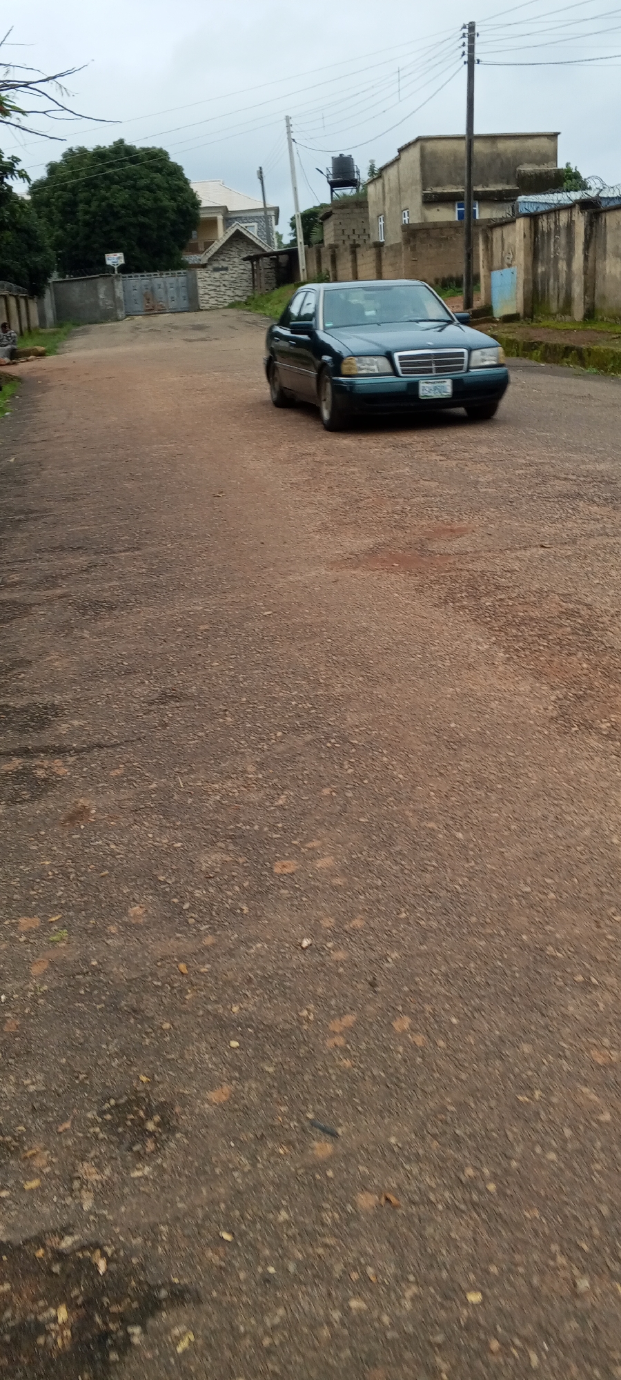 A dark green Mercedes-Benz C-Class sedan is parked on a dirt road in Bahit, Nigeria. The car is facing the viewer, with its headlights on. The road is unpaved and appears to be in a residential area. In the background, there are several houses, a large tree, and utility poles with wires. The sky is overcast. A person is sitting on the left side of the road in the foreground.