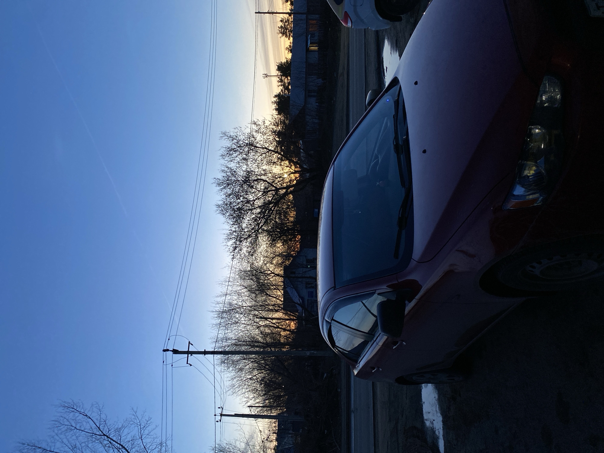 The image shows a red car parked on a street in Chaikovsky, Russia. The location is likely residential, with houses and trees visible in the background. Power lines run along the side of the road. The sun is setting, casting a warm light on the scene. The sky is a clear blue, and the air is still. There are no people visible in the image.