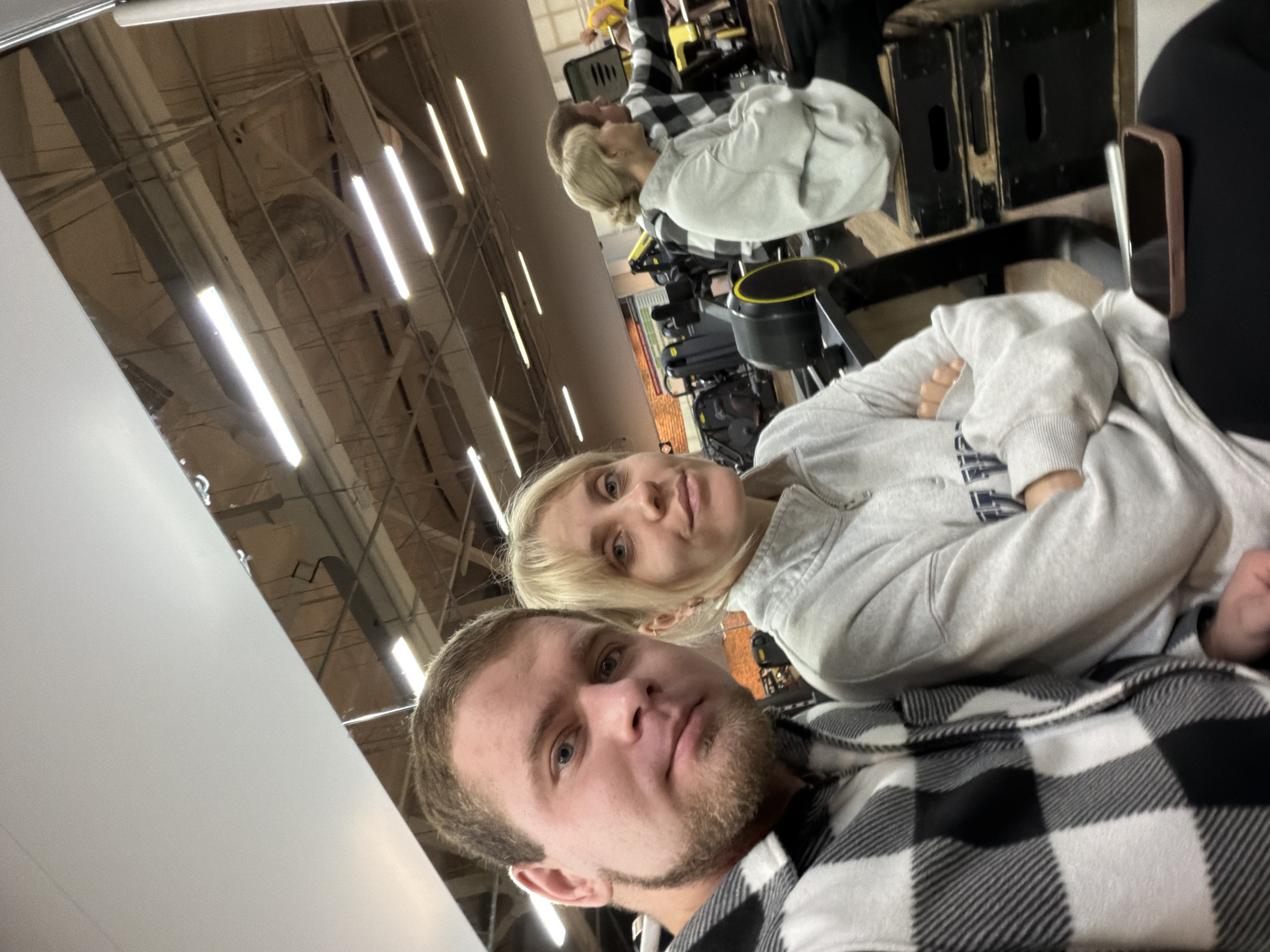 The image shows two individuals in what appears to be a gym or fitness center. A man with short hair and a beard is in the foreground, wearing a black and white checkered shirt. Behind him and slightly to the side is a woman with blonde hair, wearing a light gray hoodie. Both appear to be looking at the camera.
Gym equipment is visible in the background, including rowing machines and other exercise apparatus. The environment is illuminated with fluorescent lights, suggesting an indoor setting. The location is described as Chaikovsky, Russia.
