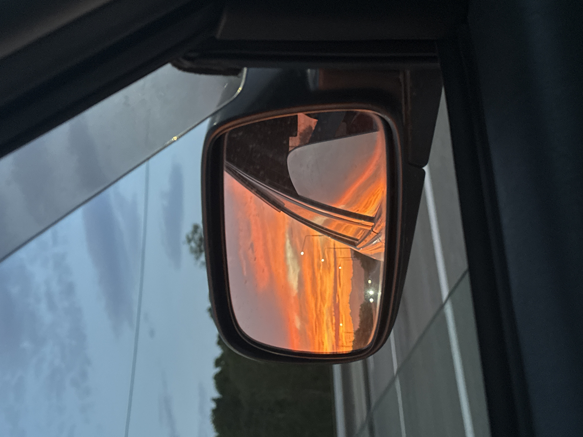 The image depicts a side-view mirror of a car, reflecting a dramatic sunset. The sky in the reflection is ablaze with vibrant shades of orange, red, and yellow, with streaks of darker clouds. Distant mountains are silhouetted against the horizon. Streetlights are visible in the reflection, suggesting the car is on or near a road. The foreground of the image shows the interior of the car, including a portion of the car door and window. The environment outside the mirror shows a road with white lines and trees along the side. There is no specific indication of the location being Yelizovo, Russia.