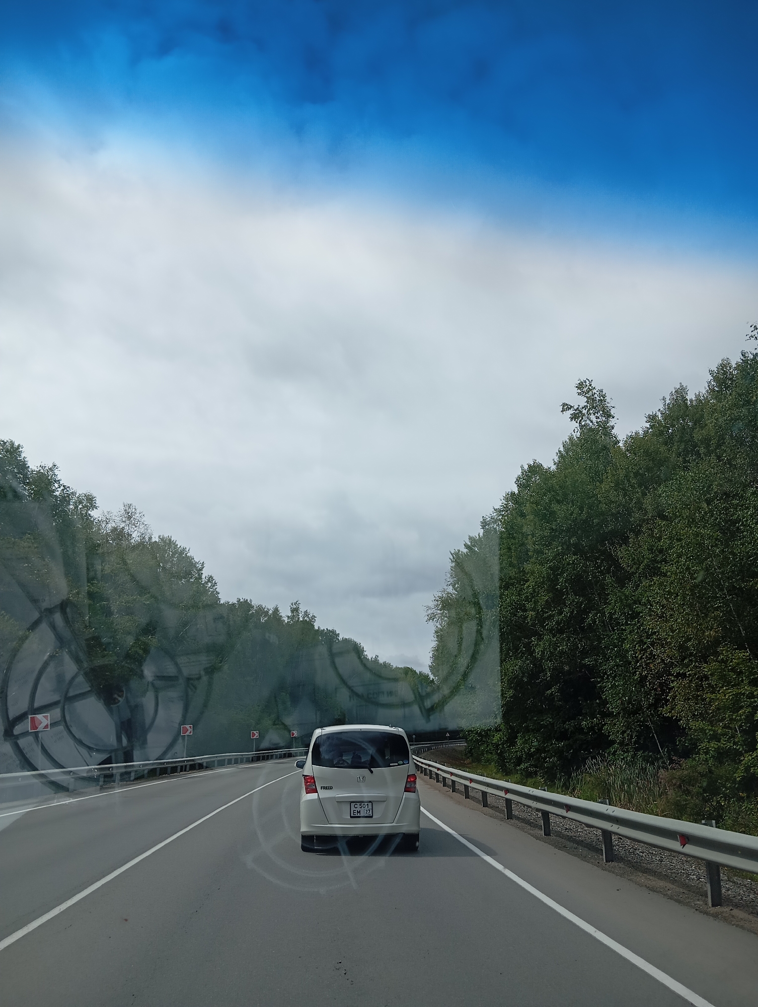The image depicts a view from inside a vehicle on a paved road, likely a highway, near Mayak, Russia. The road is divided by a white line, and there are guardrails on the right side. Dense green trees line the right side of the road, and on the left, there are large, circular, sound-absorbing barriers. The sky is overcast with patches of blue sky visible. A white car is driving in front of the camera vehicle. The license plate of the car is visible and reads "C 501 EM 27". The scene suggests a typical drive through a rural or semi-rural area under cloudy conditions.