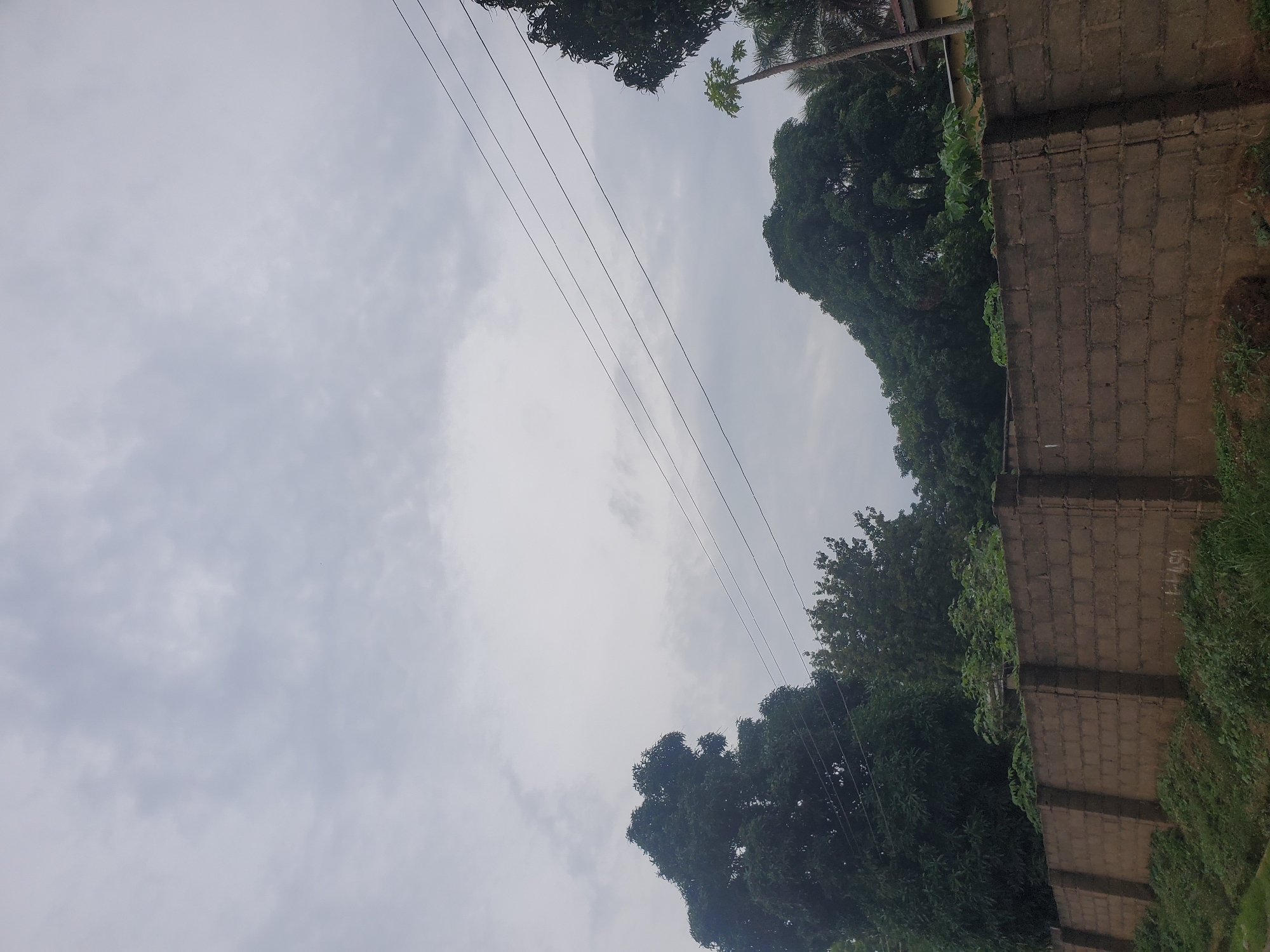 The image shows a cloudy sky over a brick wall in Jos, Nigeria. The wall is made of brown bricks and is topped with a concrete cap. There are some trees and vegetation growing behind the wall, and a few power lines are visible in the sky. A small part of a yellow building is visible to the right of the frame. The scene appears to be a residential area.