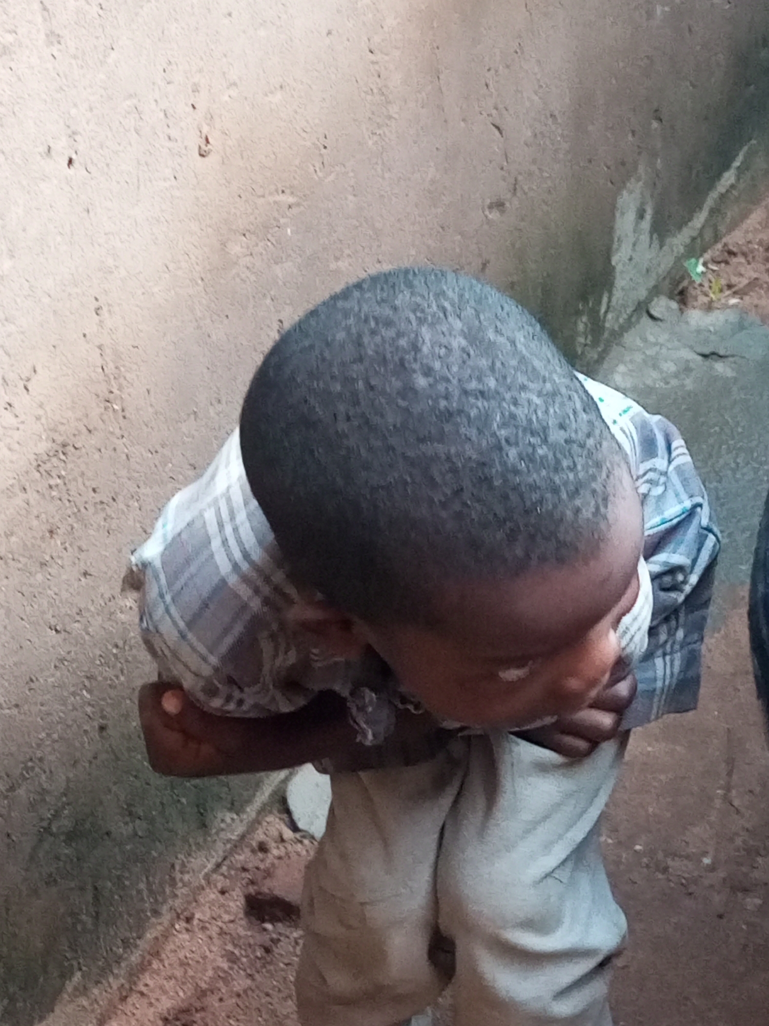 The image shows a child from a high angle view, facing away from the camera and looking down. The child has short, dark hair and is wearing a checkered shirt and light-colored pants. The child's face is partially visible, with one eye looking down and slightly to the side. The child's hands are clasped together in front of their waist. The background consists of a textured, light-colored wall on the left and a darker, rougher surface on the right, with some dirt and debris on the ground. The scene appears to be outdoors, in an urban or semi-urban environment.
