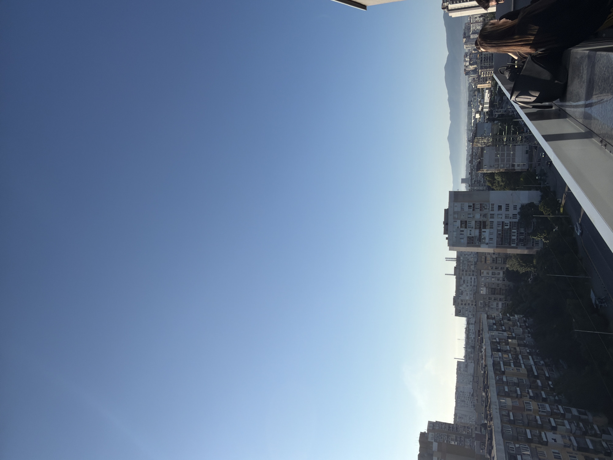 The image captures a panoramic view of Sofia, Bulgaria, from a high vantage point, likely a rooftop or balcony. The scene is bathed in the clear blue light of daytime, with a vast expanse of sky dominating the upper portion of the frame. Below, the cityscape unfolds, characterized by numerous residential buildings with varying architectural styles and heights, indicative of Soviet-era construction mixed with more modern structures. Dense foliage from trees creates a stark contrast to the urban environment. In the distance, a range of mountains is faintly visible through a haze, underscoring the geographical context of Sofia.
In the foreground, three individuals are partially visible, standing along a railing. The person on the right, a female with long brown hair, is facing away from the camera and appears to be in conversation with another individual whose back is to the viewer. A black bag is placed on the railing near them. The overall atmosphere suggests a relaxed moment of observation or social gathering in an urban setting.