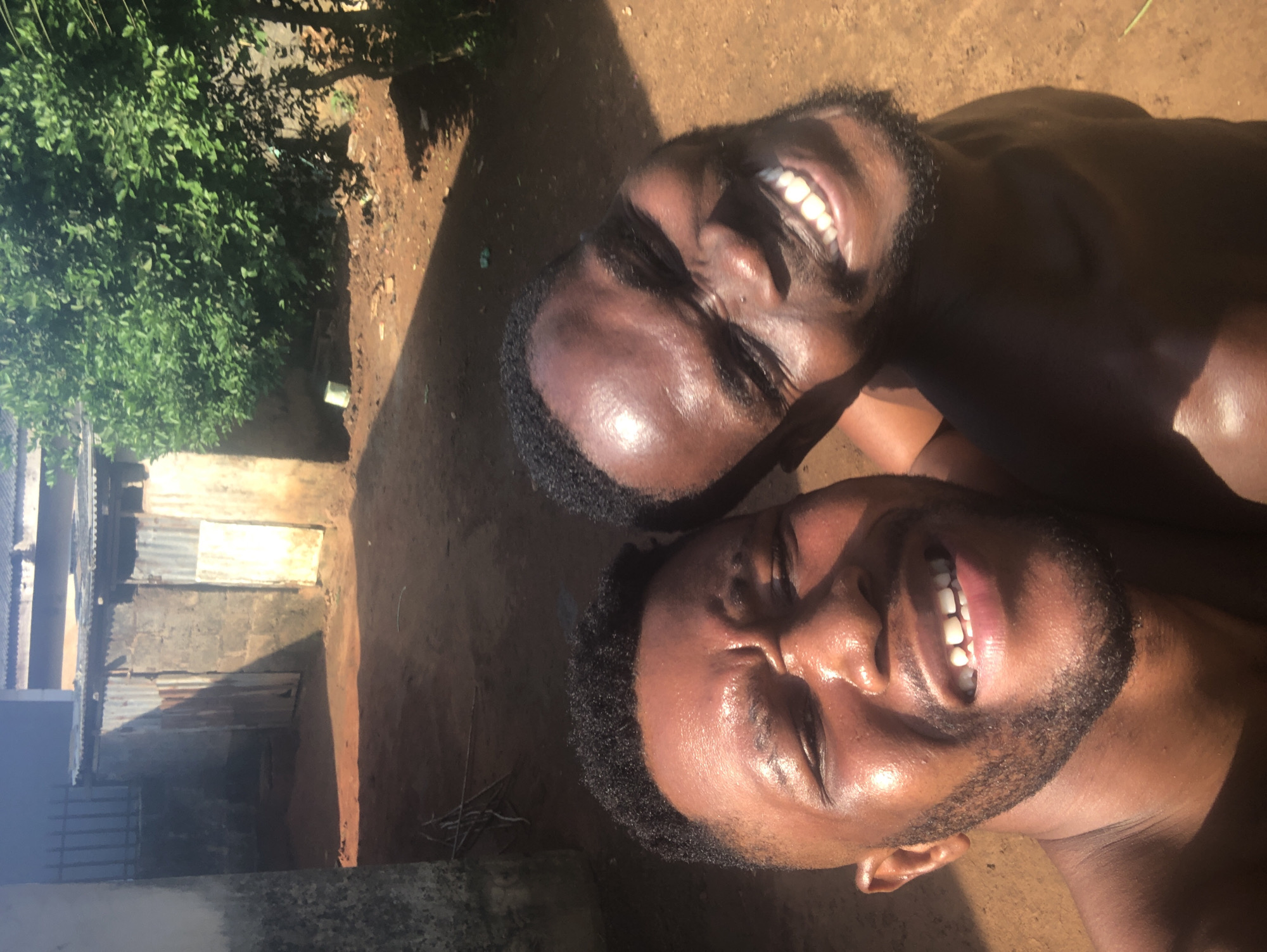 Two young men are captured in a close-up, upward-angled shot, appearing to be lying on the ground outdoors. Both have dark skin tones and short, dark hair, with prominent smiles that reveal their white teeth. Their faces are shiny, reflecting the bright, direct sunlight, suggesting warmth or recent physical activity. The man in the foreground has a neatly trimmed beard and his eyes are partially closed, possibly due to the intense sun or the expression of his wide smile. The man positioned slightly behind him also smiles broadly, looking up towards the camera.
The background features sun-baked, unpaved ground, indicating a natural or rustic outdoor setting. To the left, lush green foliage from trees or bushes is visible, contrasting with the earthy tones of the ground. Further into the background, parts of simple structures with corrugated metal roofs can be seen, hinting at a residential or village environment. The scene is brightly illuminated, with strong shadows cast, confirming a sunny daytime setting. The men's expressions convey happiness and relaxation. No visible text is present in the image.