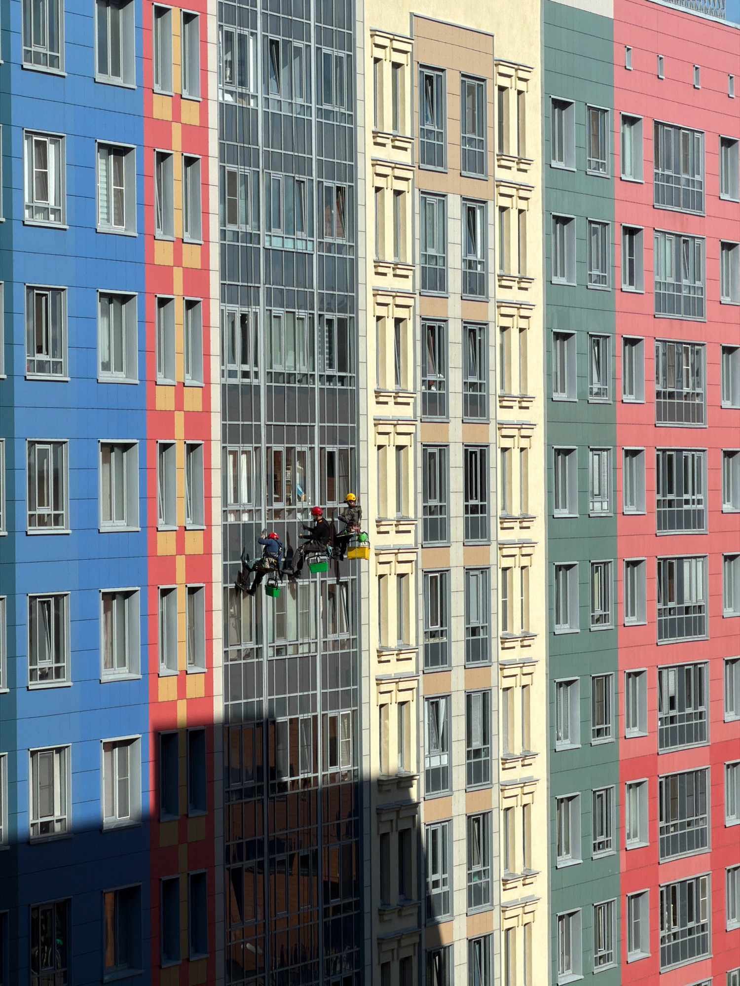 The image shows a brightly colored, modern apartment building in Saint Petersburg, Russia. The building features vertical sections of different colors, including blue, red, yellow, beige, green, and pink. The facade is composed of many windows, with some sections having a glass exterior.
On the glass facade of the building, three window washers are suspended on a platform, working at height. They are wearing protective gear, including helmets. One individual appears to be operating the cleaning equipment. The window washers are positioned on the central section of the building, which is largely glass. Their platform is equipped with buckets, one of which is yellow and the other green, likely holding cleaning supplies.
The lighting suggests it is daytime, with sunlight casting shadows on the building's facade. The windows reflect the sky and surrounding environment, adding depth to the scene. The overall impression is one of urban development and the maintenance of modern architecture.