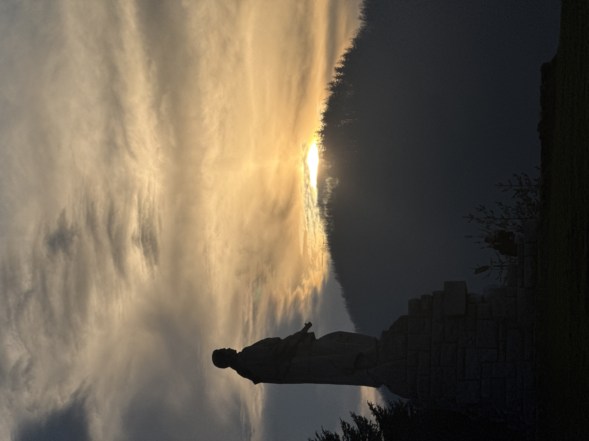 The image shows a silhouette of a statue against a bright, cloudy sky with a partially obscured sun. The statue appears to be of a man in robes, standing on a stone base. The environment around the statue is dark, suggesting a shaded or nighttime setting. The location is Etropole, Bulgaria, which is known for its historical landmarks and monasteries.