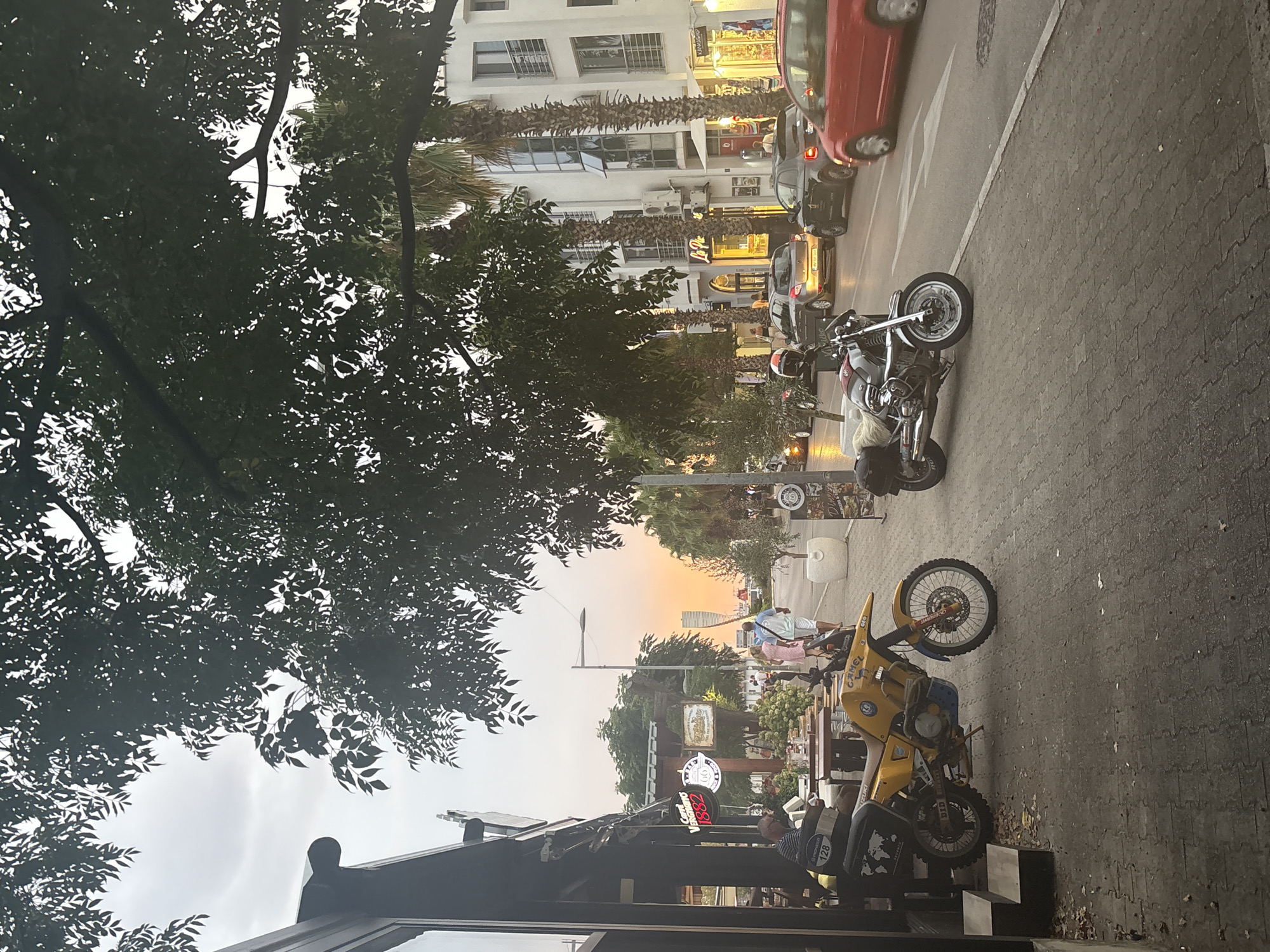This is a photograph of a street scene in Bar, Montenegro, during a sunset. The image features a sidewalk with a yellow motorcycle parked on the left and a silver motorcycle parked on the right. A building with shops and restaurants is on the left side of the street, with outdoor seating areas. A few people are visible walking on the sidewalk and sitting at tables. Several cars are driving on the street, including a red car in the foreground. Palm trees and leafy trees line the street, and the sky is colored with the warm hues of the sunset. The overall impression is that of a lively street in a coastal town during the evening.