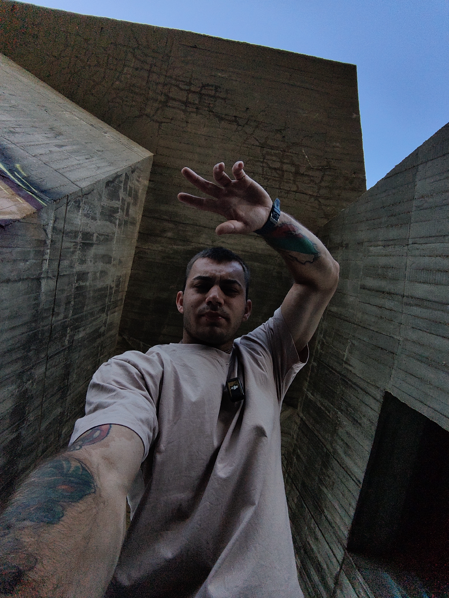 A low-angle, wide shot captures a man standing between two brutalist concrete structures, possibly part of an abandoned Soviet-era military facility, under a clear blue sky. The man, with short dark hair and a slight beard, is wearing a light beige t-shirt and has a small black device clipped to his shirt. He has tattoos on his left arm and shoulder, and his right arm is raised with his hand covering his face. The concrete surfaces show signs of age, with visible lines from the forms used in their construction and some graffiti on the left structure. The man's expression is obscured, but his raised arm suggests a pose or interaction with the camera. The scene appears to be an outdoor urban environment in Varna, Bulgaria, characterized by its imposing concrete architecture and open sky.