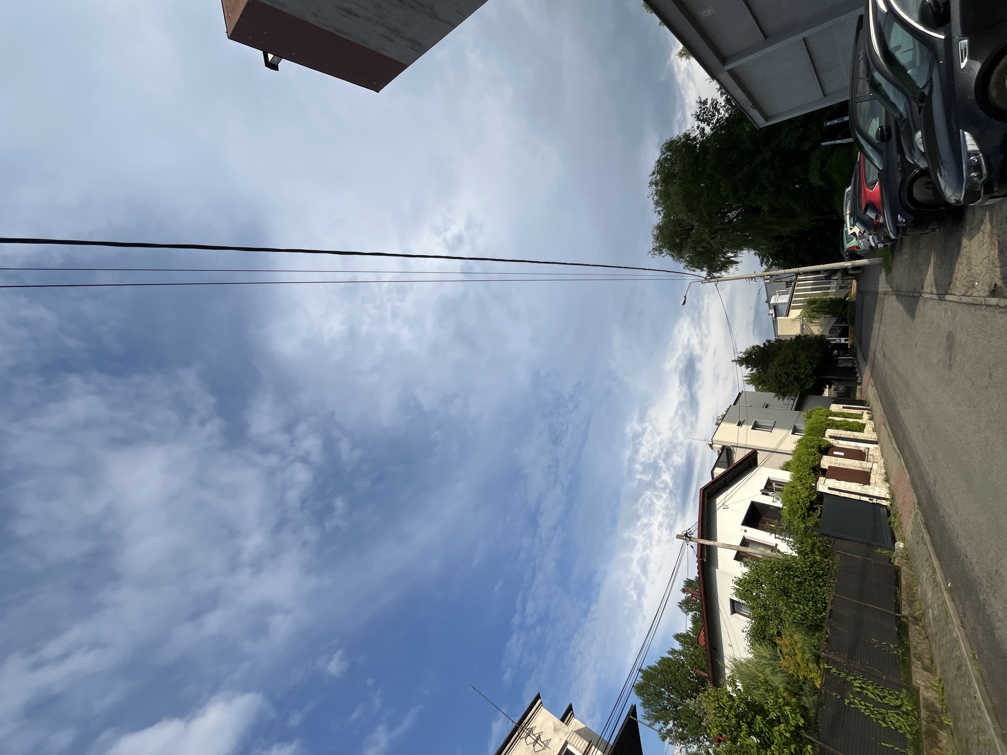 The image captures an outdoor scene, looking upwards towards a blue sky filled with scattered white clouds of varying density. Utility lines stretch horizontally across the upper part of the frame.
On the right side of the image, a paved residential street is visible, with a curb or sidewalk running alongside it. Several cars, including dark, red, and white vehicles, are parked bumper-to-bumper along the street. Behind the cars and street are multiple houses, predominantly light-colored with dark-colored fences and gates. Lush green trees and bushes line the properties, providing significant foliage. A utility pole with an attached street lamp stands prominently near the houses. On the far left, the corner of a building, possibly a multi-story structure with a flat top, is partially visible. Towards the bottom left, another building's top edge and an antenna are present.
The scene depicts a quiet street on a bright, cloudy day. No specific activity or event is taking place, and no people are visible. There is no discernible text within the image.