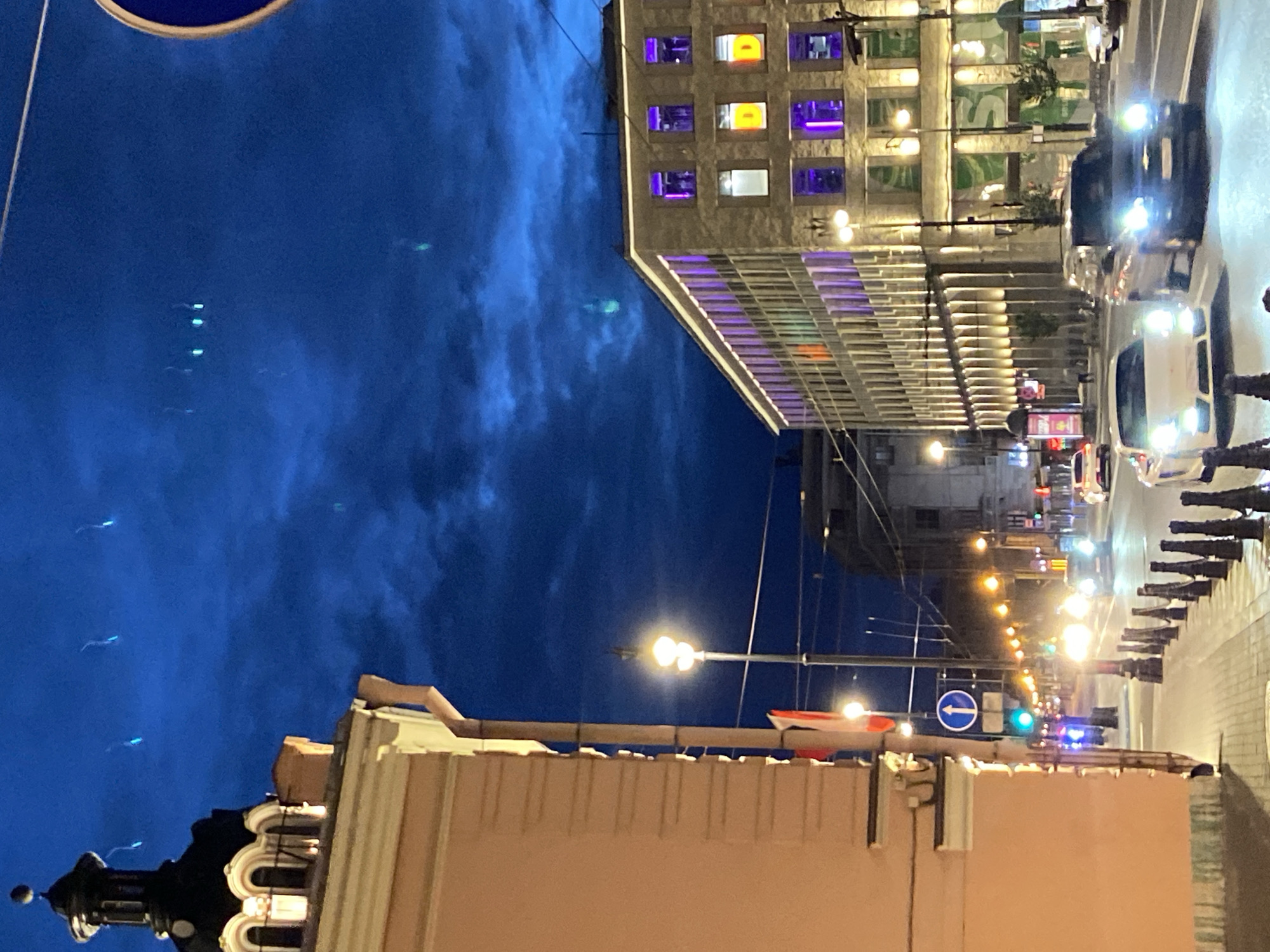This image depicts a street scene in Saint Petersburg, Russia, at dusk or night. The sky is a deep blue, with streaks of light, possibly from fireworks or flares, visible in the upper portion of the frame.
On the left, a building with an ornate dome and arched windows is partially visible. Adjacent to it, a light-pink building occupies the foreground, with a downspout running along its side.
The street is illuminated by streetlights and the lights from buildings. Several cars are visible on the road, including a white sports car and a dark SUV. Traffic lights are also present, with a green light visible. A traffic sign with a blue circle and a white arrow pointing upwards is also seen.
On the right side of the street, a modern building with numerous windows is illuminated with purple and orange lights, suggesting it is an office building or a commercial establishment. The lower floors of this building appear to be shops or restaurants, with some visible signage.
The sidewalk is lined with bollards, and there are street lamps with multiple lights. The overall impression is of a well-lit urban environment, likely a commercial or residential area.