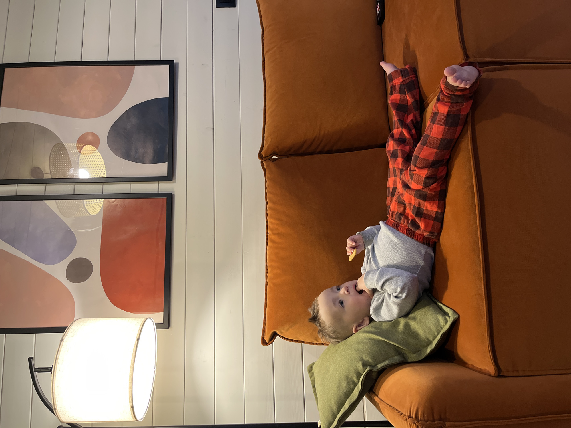 An indoor scene features a young child lying on their back on a rust-orange couch, with their legs elevated and feet pointing upwards. The child is wearing red and black plaid pants and a light grey long-sleeved shirt. A small, olive-green throw pillow supports their head and upper back. The child is holding a light-colored object, possibly a snack, to their mouth with one hand, while the other hand is also near their face. Their expression is attentive and slightly curious.
The couch is a modular design with plush, soft cushions, upholstered in a warm, rust-orange fabric. Behind the couch, a wall with vertical white wood paneling serves as the backdrop. Two large, rectangular framed art prints hang vertically on the wall. Both prints feature abstract, organic shapes in a palette of muted colors, including pink, grey, dark blue, and red. The glow from a modern floor lamp, with a wide, drum-shaped shade, illuminates the lower left portion of the image and is reflected in the glass of the picture frames. The overall setting appears to be a cozy living space, lit by artificial light. No visible text is present in the image.