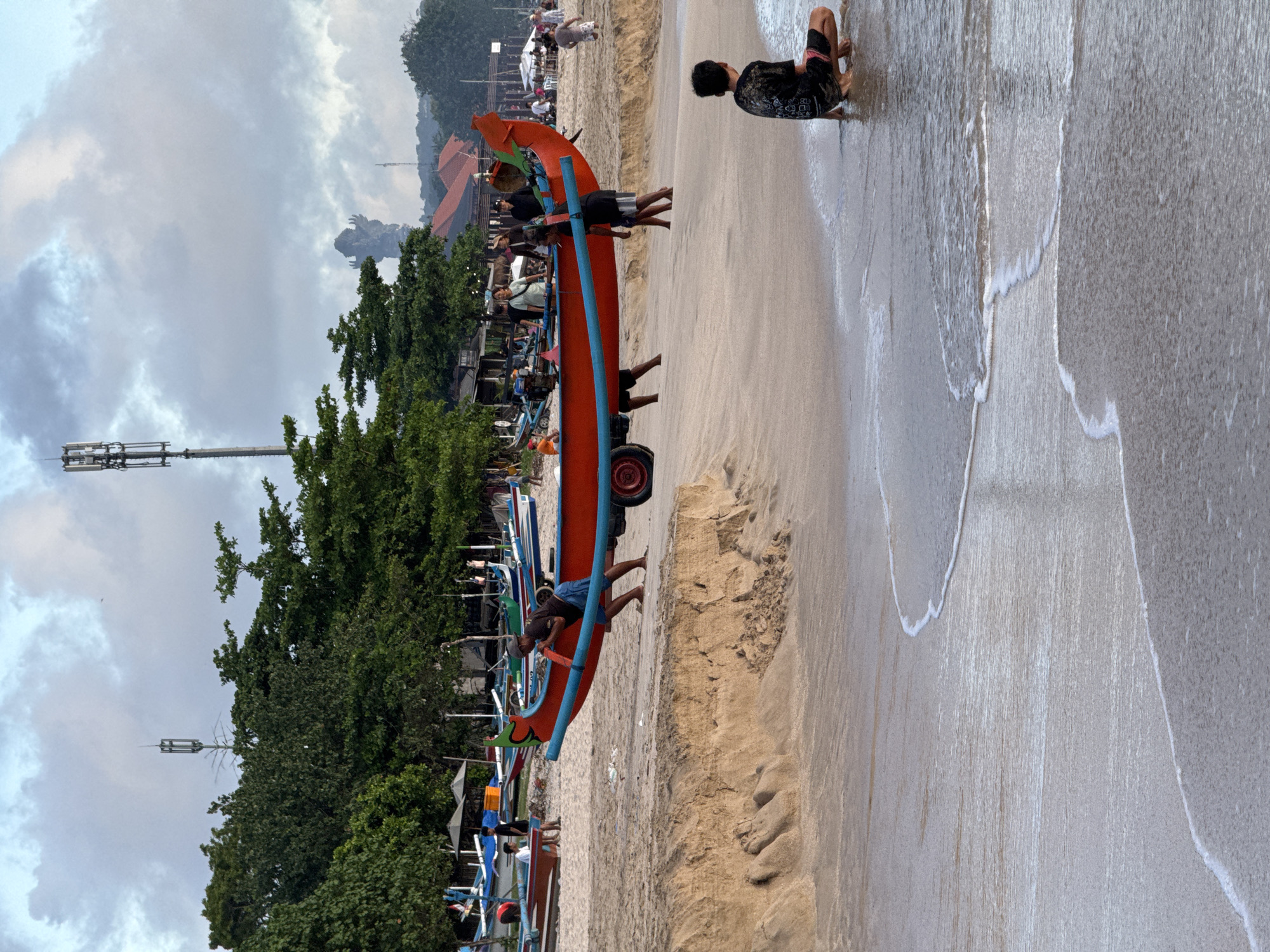 The image shows a beach scene in Kuta, Indonesia. A group of men are pushing a large orange outrigger canoe on wheels along the sand near the water's edge. Another person is kneeling at the edge of the water. The beach is tan, wet from the surf, and has numerous boats docked in the background next to trees and buildings. Above, there are gray clouds in the sky. Visible in the distance are structures that appear to be cellular towers.