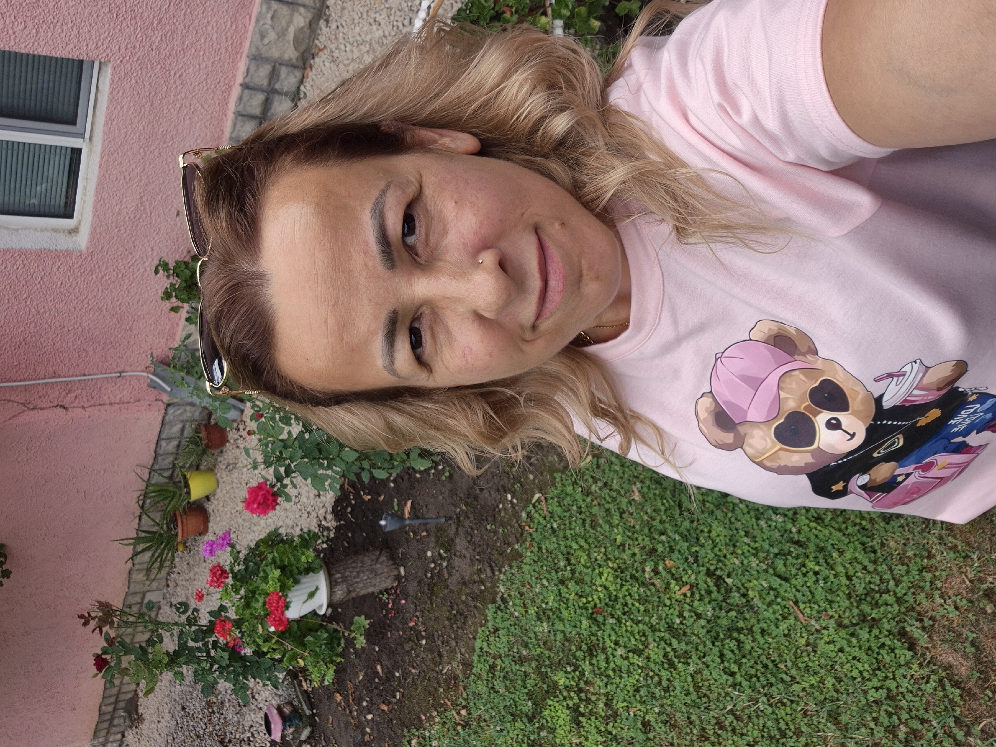 A blonde woman with light skin and a pink t-shirt featuring a teddy bear graphic poses for a selfie in an outdoor setting. She is wearing sunglasses perched on her head and has a nose ring. The background shows a pink-walled building with a window, a gravel path, and a garden with various plants, including roses and succulents in pots. A patch of green grass is visible in the foreground.
