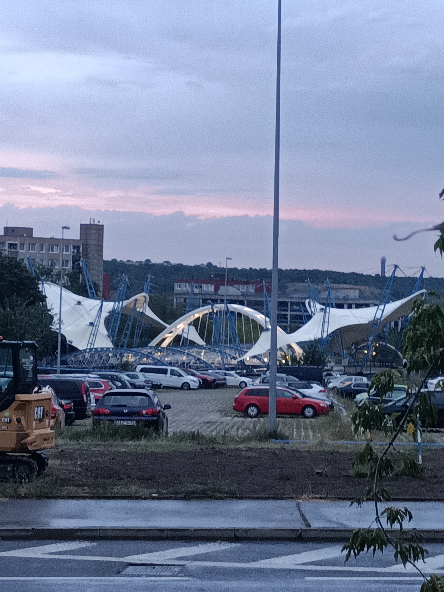 The image captures an outdoor scene with a large, white, tent-like structure supported by blue scaffolding. In the foreground, a yellow construction vehicle with "CAT" branding is visible on the left. A parking lot filled with various cars is present behind the construction vehicle, stretching towards the tent structure. In the background, several multi-story buildings and a hill covered with trees are visible under a twilight sky with soft pink and purple hues. The overall impression is that of an urban or semi-urban environment at dusk. Based on the visual cues and architectural style, this scene is likely located in Prague, Czechia, possibly near a venue or event space given the distinctive tent structure.