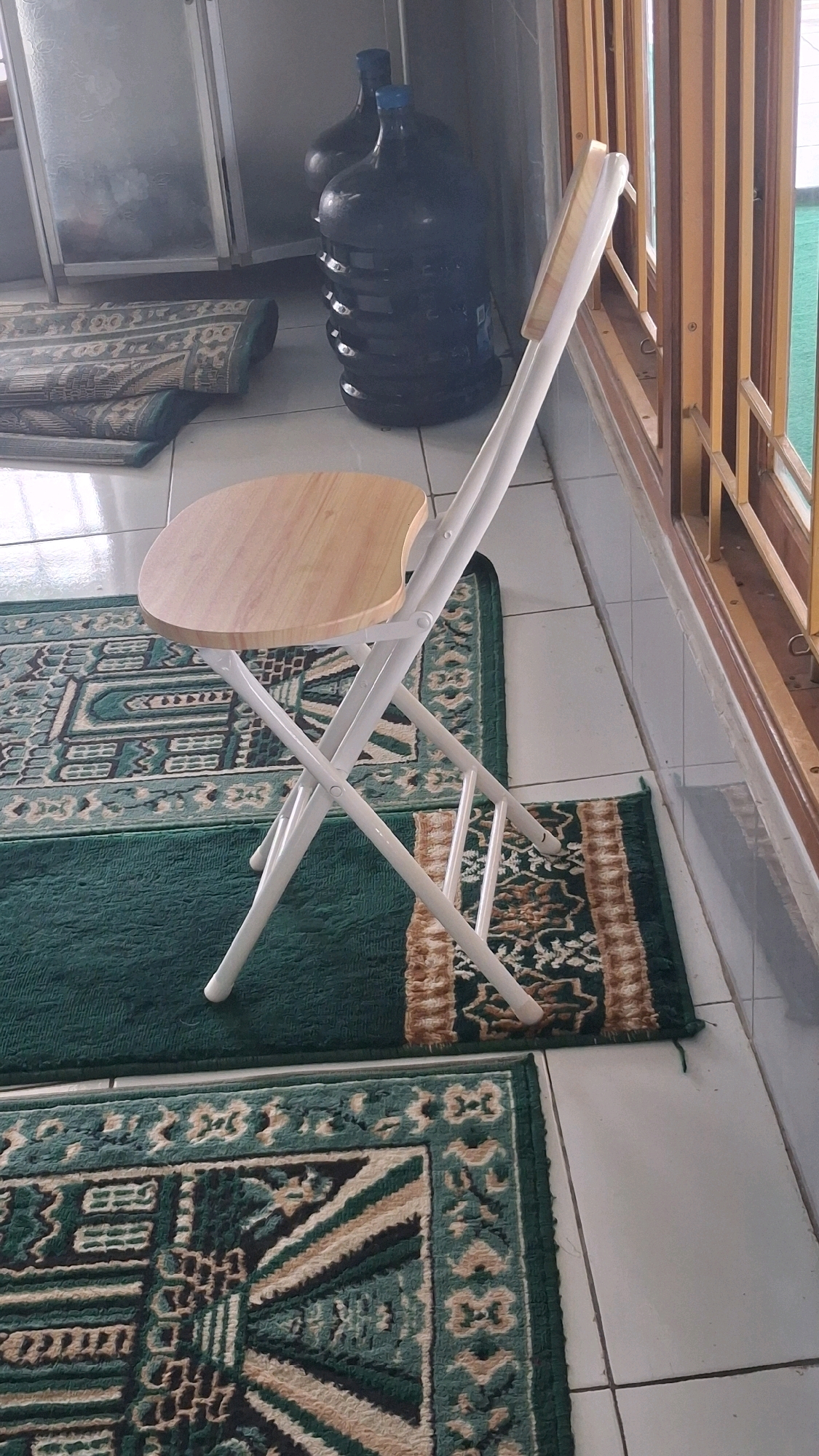 The image depicts an indoor scene, likely a home or a communal space, with elements suggesting a religious context. A white foldable chair with a light-colored wooden seat is positioned centrally. The chair is set on top of two prayer rugs, one primarily green and the other with a patterned design in green, beige, and brown. In the background, two large, dark blue water jugs are visible next to a cabinet with reflective doors. A window with wooden-slatted blinds is on the right side of the frame, revealing a glimpse of greenery outside. The floor is tiled in a light, neutral color. The overall impression is one of a simple, functional setting. The presence of prayer rugs and water jugs commonly used for religious purposes suggests a connection to Islamic practice, and therefore, a possible location in a Muslim-majority area like Giriwarno, Indonesia.