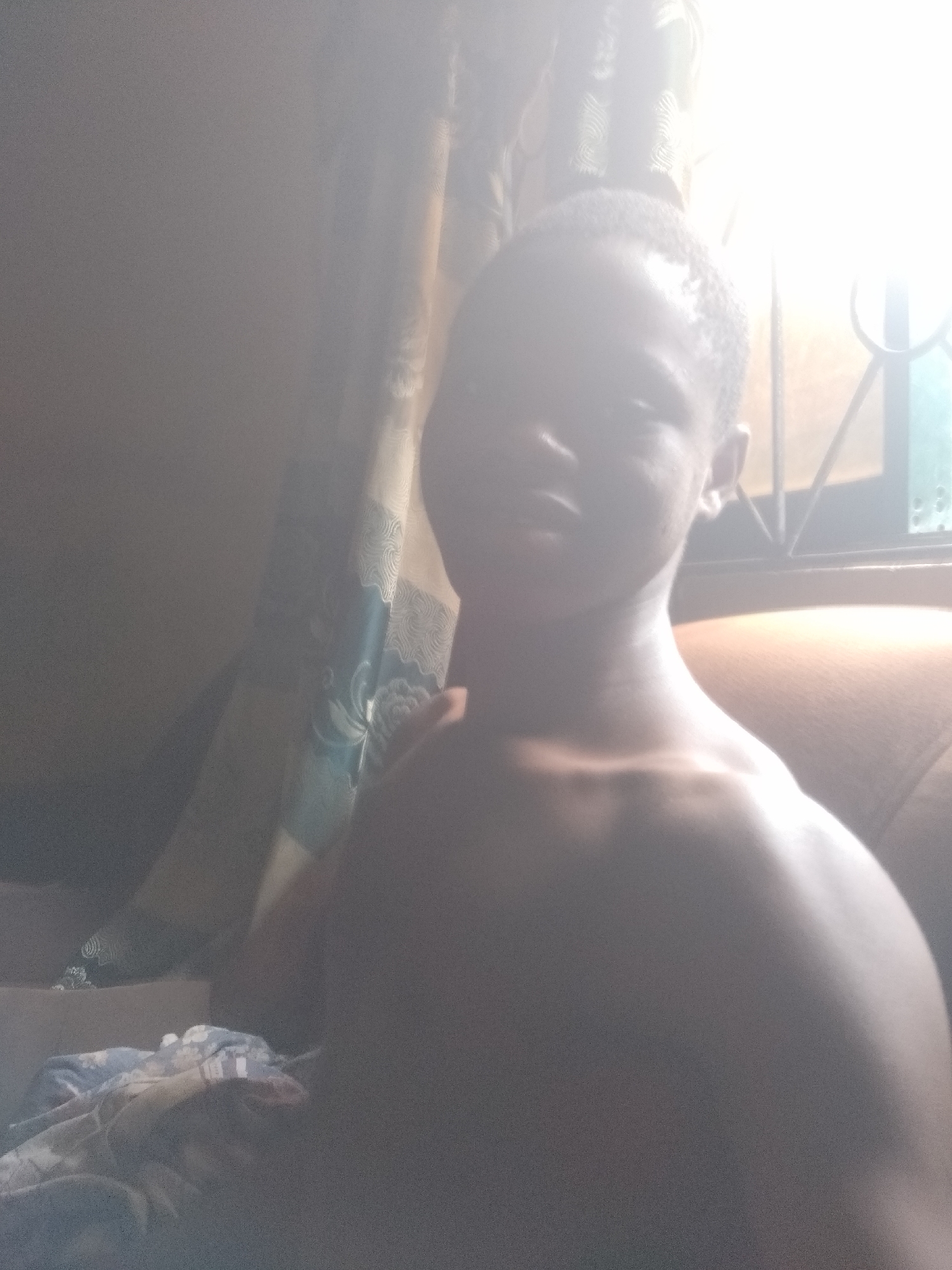The image features a young person, likely male, captured in a close-up portrait from the chest up. The subject appears to be bare-chested, facing forward towards the camera. Intense backlighting originating from a window behind and to the right of the subject causes severe overexposure across the majority of the image, obscuring most facial features and creating a bright, hazy glow.
In the background, a patterned curtain, featuring blue, teal, and white swirling motifs, is partially visible. To the right, a window with discernible metal bars or a grid allows bright sunlight to flood the scene. Faintly visible behind the subject's right shoulder is what appears to be a brown cushion or piece of upholstered furniture. In the lower-left foreground, out-of-focus fabric with a blue and white pattern, possibly bedding or clothing, is present, resting on an indistinct object that resembles a box.
The extreme brightness indicates the photograph was taken during daytime, possibly with direct sunlight entering the room. The setting is an indoor environment, likely a bedroom or living space, in Shangisha, Nigeria. The primary activity is the subject posing for the camera. There is no visible text in the image.