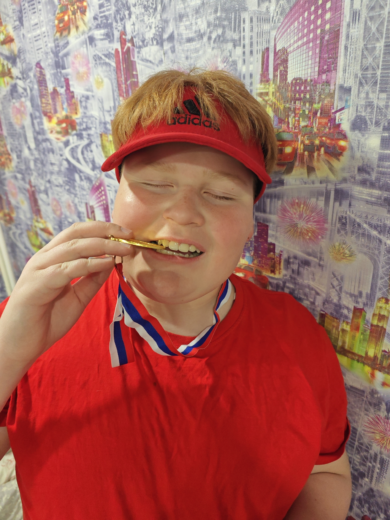 A young Caucasian male with ginger hair is biting a gold medal with a blue, white, and red ribbon. He is wearing a red T-shirt and a red Adidas visor. The background features wallpaper depicting a stylized cityscape. The location in the city of Markovskii, Russia, is unconfirmed.
