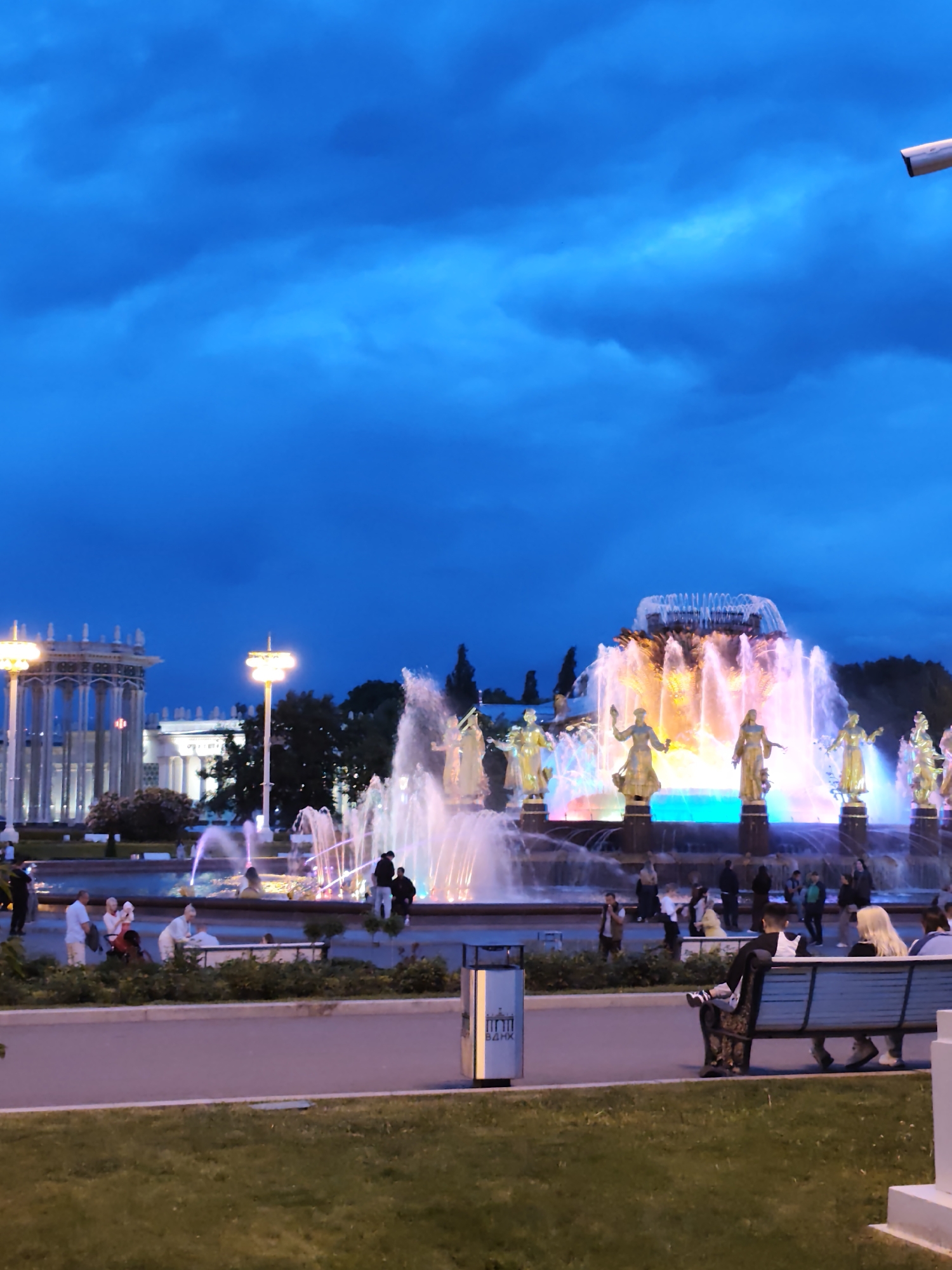 At night, Moscow, Russia, features a fountain complex adorned with gold statues. The fountain is illuminated with vibrant colors. People are seen strolling and relaxing on benches, while others gather near the fountains. Nearby, a gazebo is visible in the background. The sky is dark blue. A security camera is mounted at the top right of the image.
