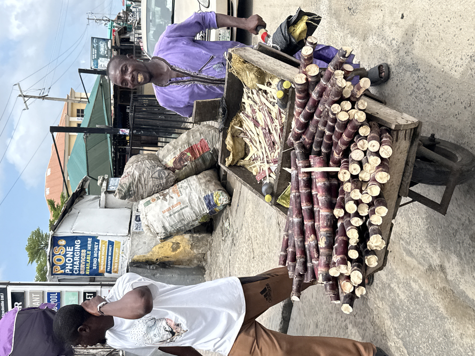 The image depicts an outdoor scene in Ado, Nigeria, featuring a man selling sugarcane from a handcart on a city street. The vendor, dressed in a purple shirt with decorative trim, smiles at the camera. The cart is laden with bundles of sugarcane, along with what appear to be bags of flour and other goods. Nearby, a younger man in a white t-shirt and brown pants is looking at his phone. In the background, there is a small business offering POS (Point of Sale) phone charging services and money transfer options, as indicated by the signs, as well as a car wash/bar. The environment is urban, with buildings and overhead power lines visible.
