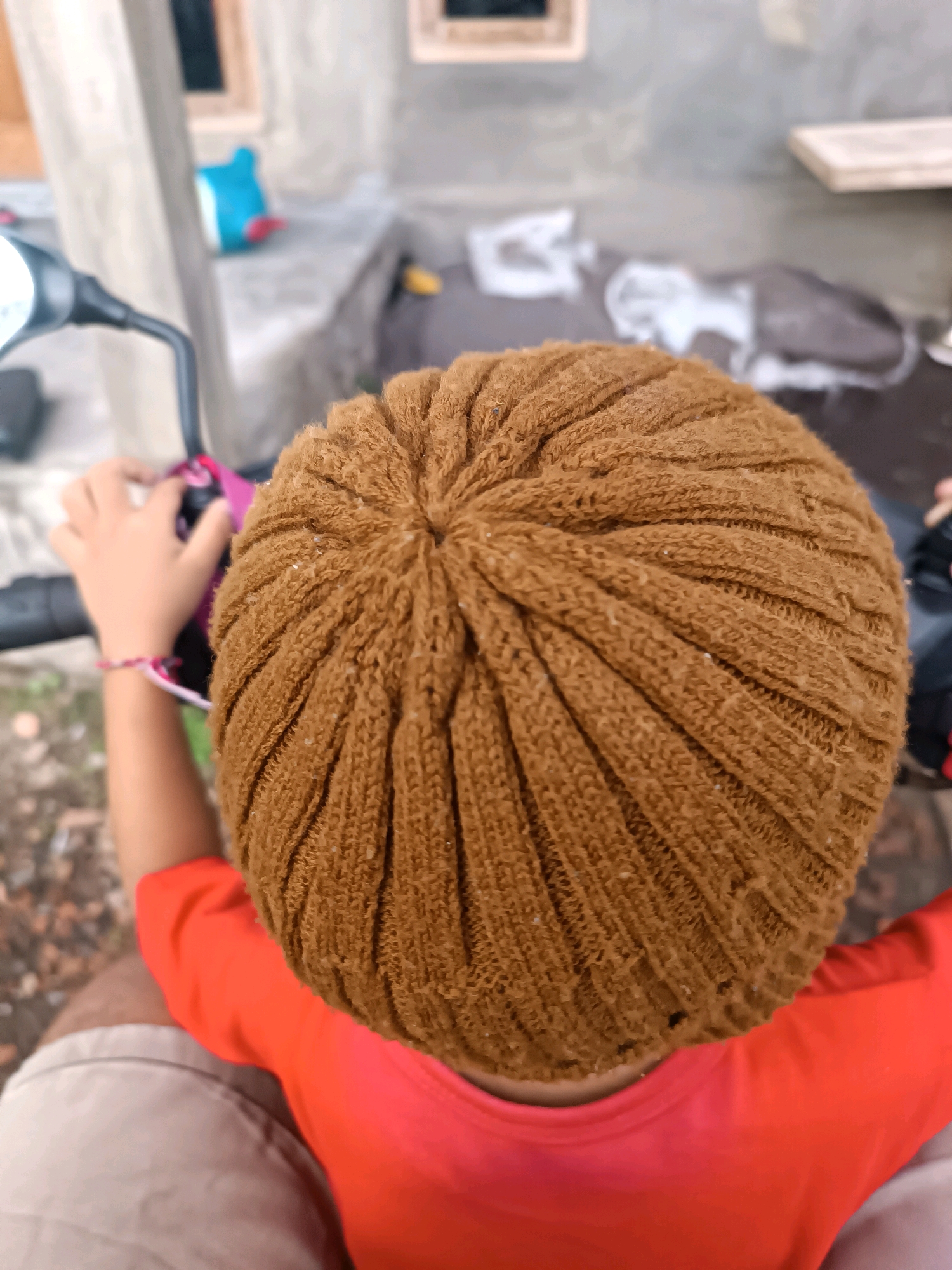 An overhead, eye-level shot shows the top of a person's head covered by a brown knitted beanie. The beanie has vertical ribbed texture and is slightly fuzzy. A child's hand wearing a thin pink and white bracelet is visible on the left, gripping the handle of a black object, possibly a motorcycle handlebar, with a portion of a silver mirror to its left. The child is wearing a bright orange t-shirt, and beige shorts are visible at the bottom of the frame. The background is out of focus and appears to be an outdoor environment, possibly near a building with concrete elements and some debris on the ground. The location is the city of Tabanan, Indonesia.