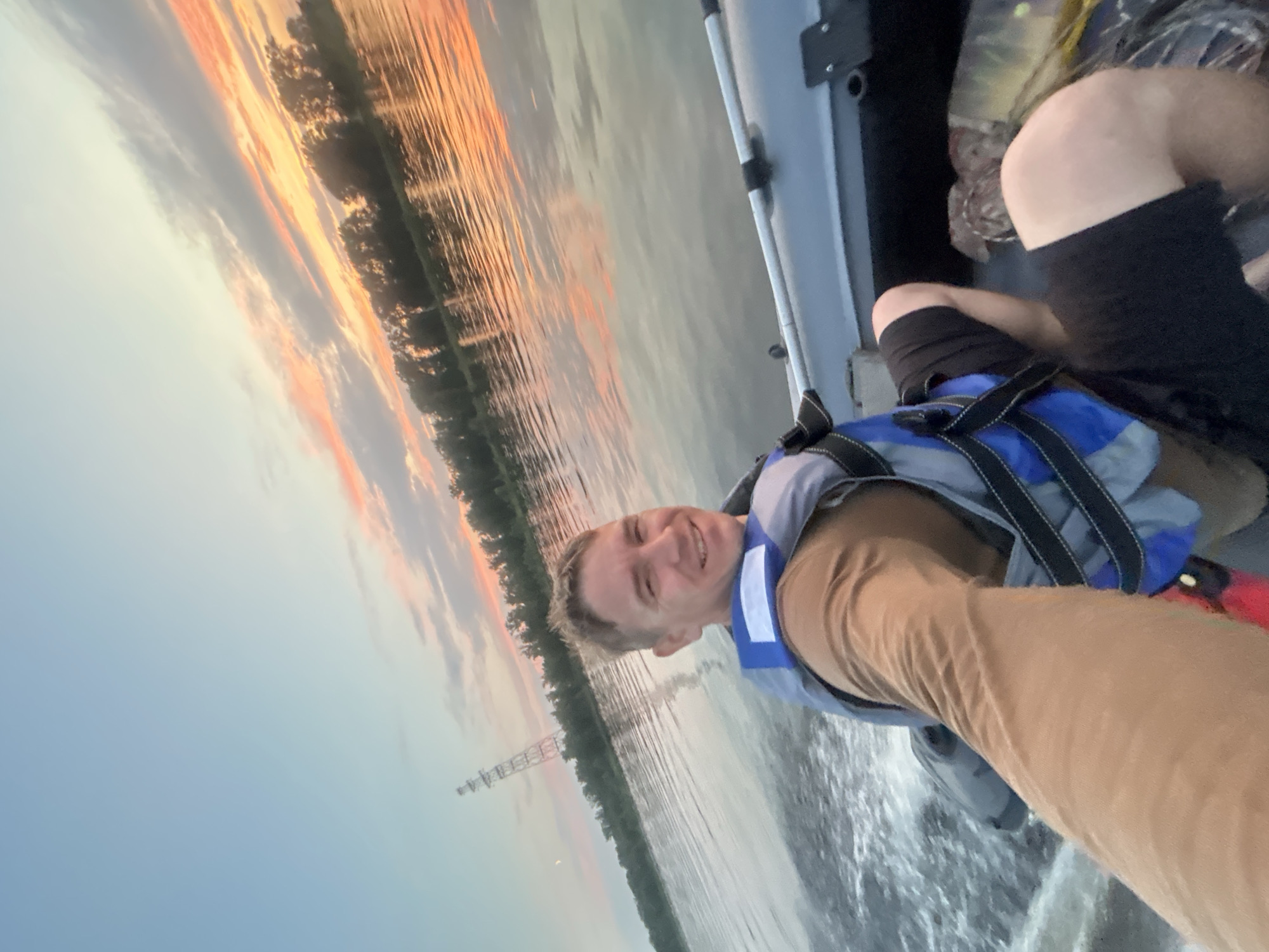 A man wearing a brown shirt and a blue life vest is taking a selfie while riding in a small gray inflatable boat on a body of water. The boat is moving, creating a wake. The man is smiling and looking at the camera. In the background, there is a shoreline with trees and a tall, lattice-like tower structure. The sky is a mix of blue with streaks of orange and pink from a sunset or sunrise. The water reflects the sky and trees. The scene appears to be outdoors on a river or lake.