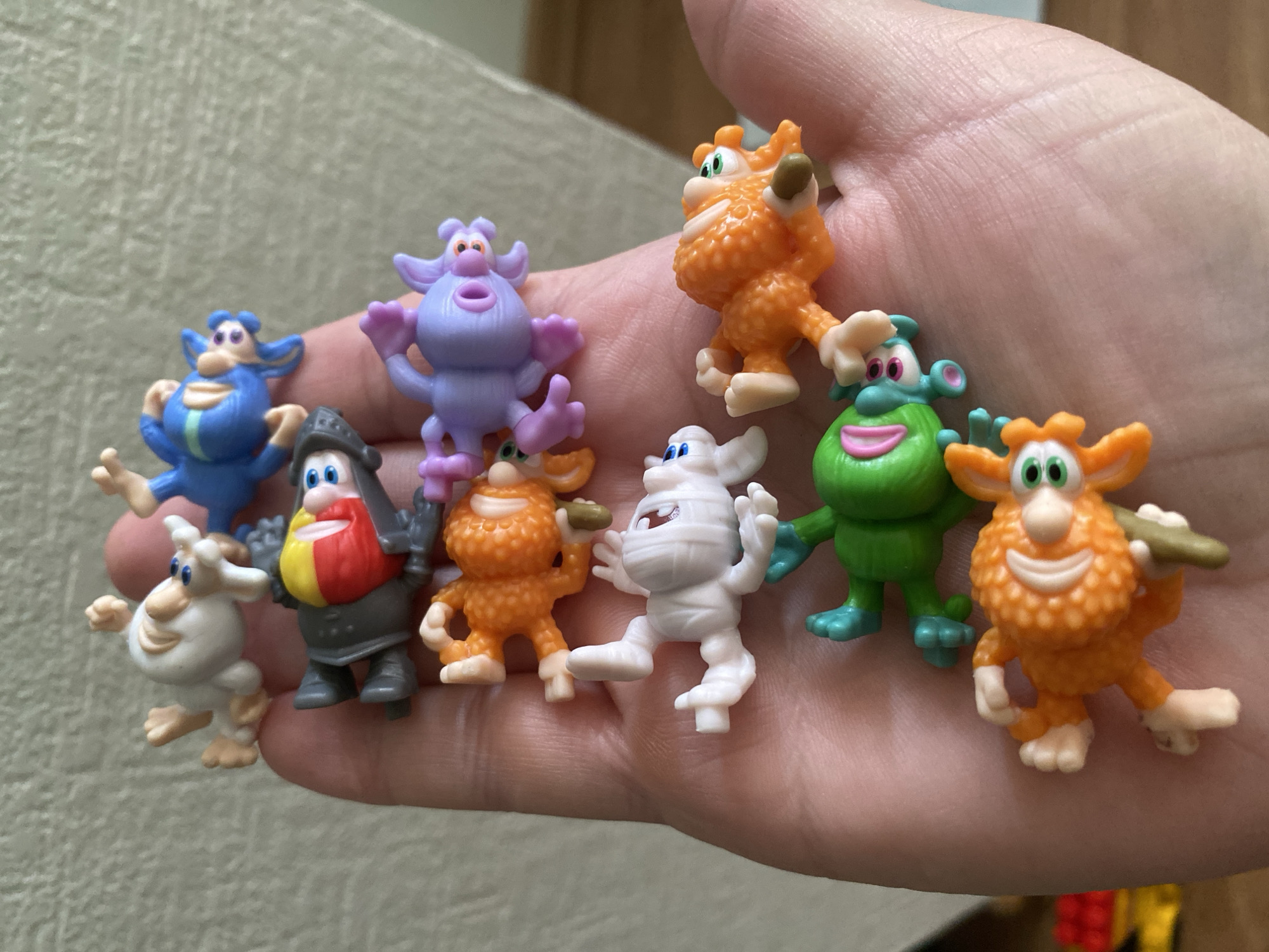 A person's hand holds ten small, colorful, plastic figurines that resemble cartoon monsters. The figurines are arranged in a cluster on the palm. The background is a soft, light-brown texture. There's no information to suggest the location is in Sochi, Russia.