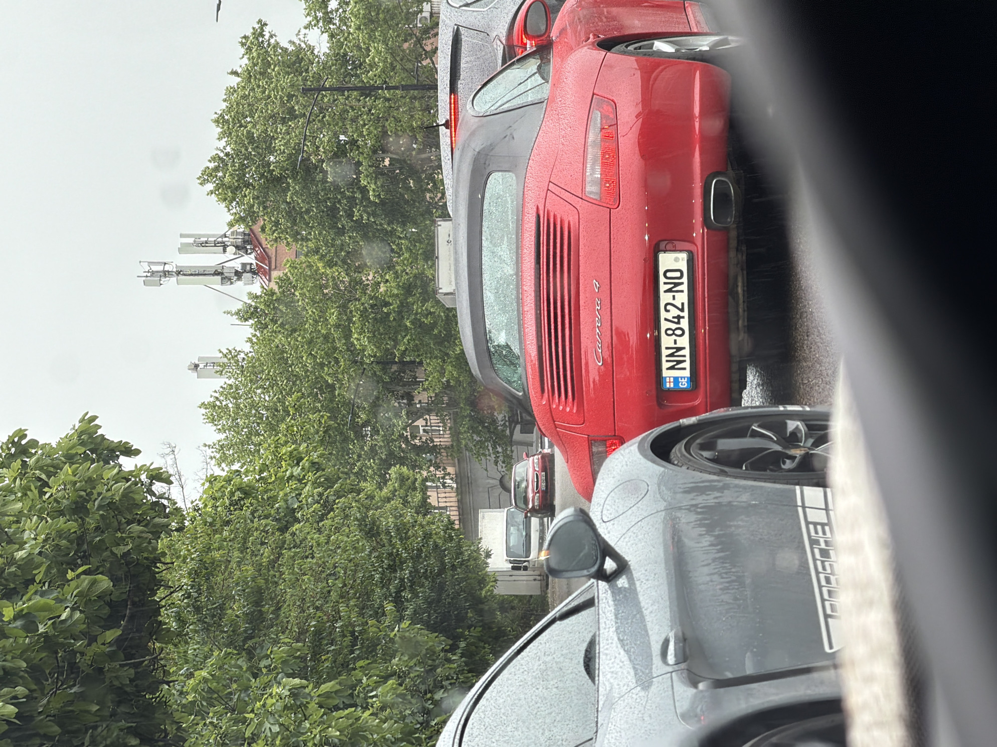 The image shows a street scene in Tbilisi, Georgia, likely taken from inside a car during a rainy day. A red Porsche Carrera 4 is directly in front, its license plate reads "NN-842-NO" and has the Georgian flag. A grey Porsche with the word "Porsche" vertically printed on the side is to the right, next to the window of the car from which the photo was taken. Other cars are visible in the distance between the trees. The left side of the image is dominated by green trees. In the distance, antennas and a building can be seen.