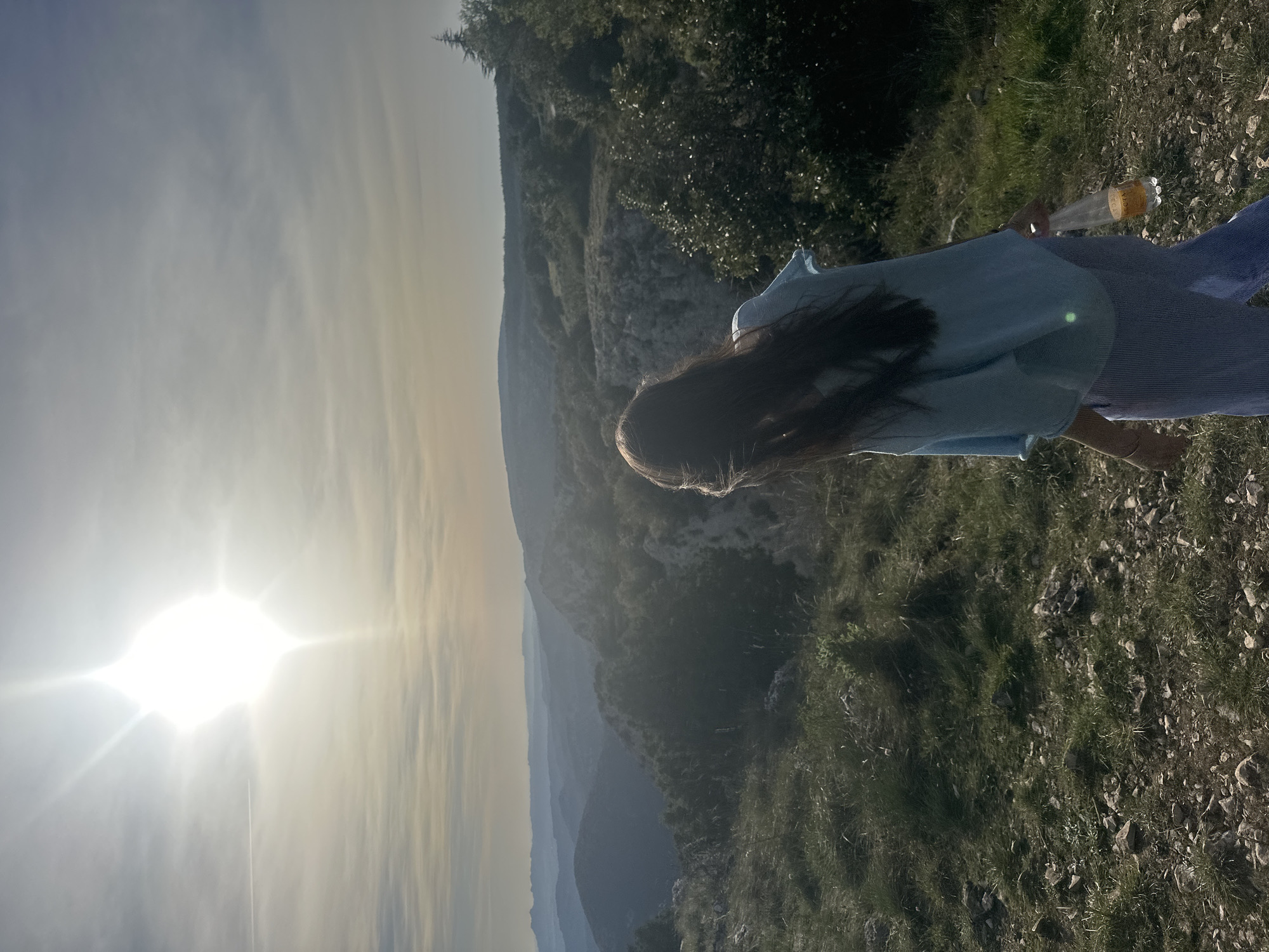 A woman with long dark hair stands on rocky, grassy terrain, possibly a cliffside or elevated area, near Bonnieux, France. She is wearing a light blue top and blue pants. In her hand, she is holding a plastic bottle containing a yellow liquid. The background shows a mountainous landscape under a bright sky, possibly during sunrise or sunset due to the angle and intensity of the sunlight. The environment consists of green vegetation and rocky surfaces.
